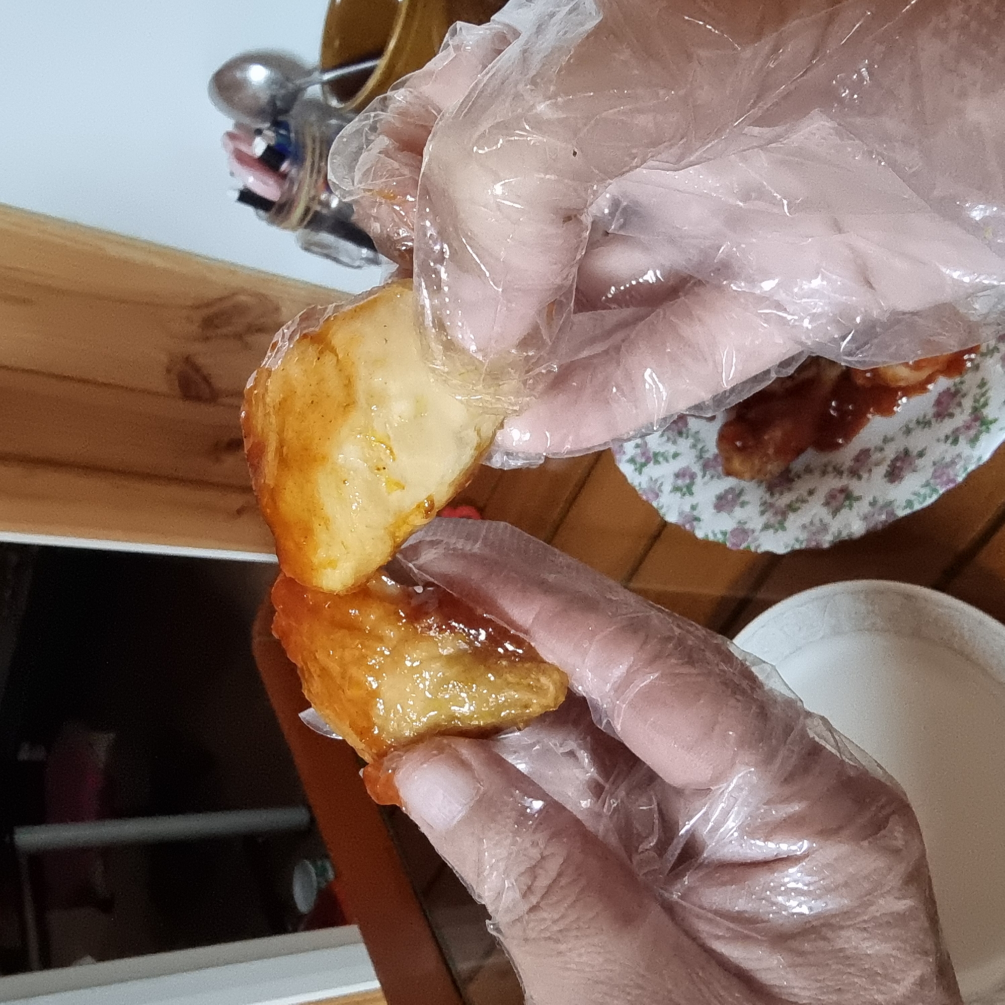 Two gloved hands hold pieces of fried food coated in a shiny glaze. The food appears to be chicken wings or tenders. In the foreground, a small plate with a floral pattern holds a piece of similar glazed food. The background shows a wooden table and a blurred interior setting. The image was taken in Dunnae-myeon, South Korea.