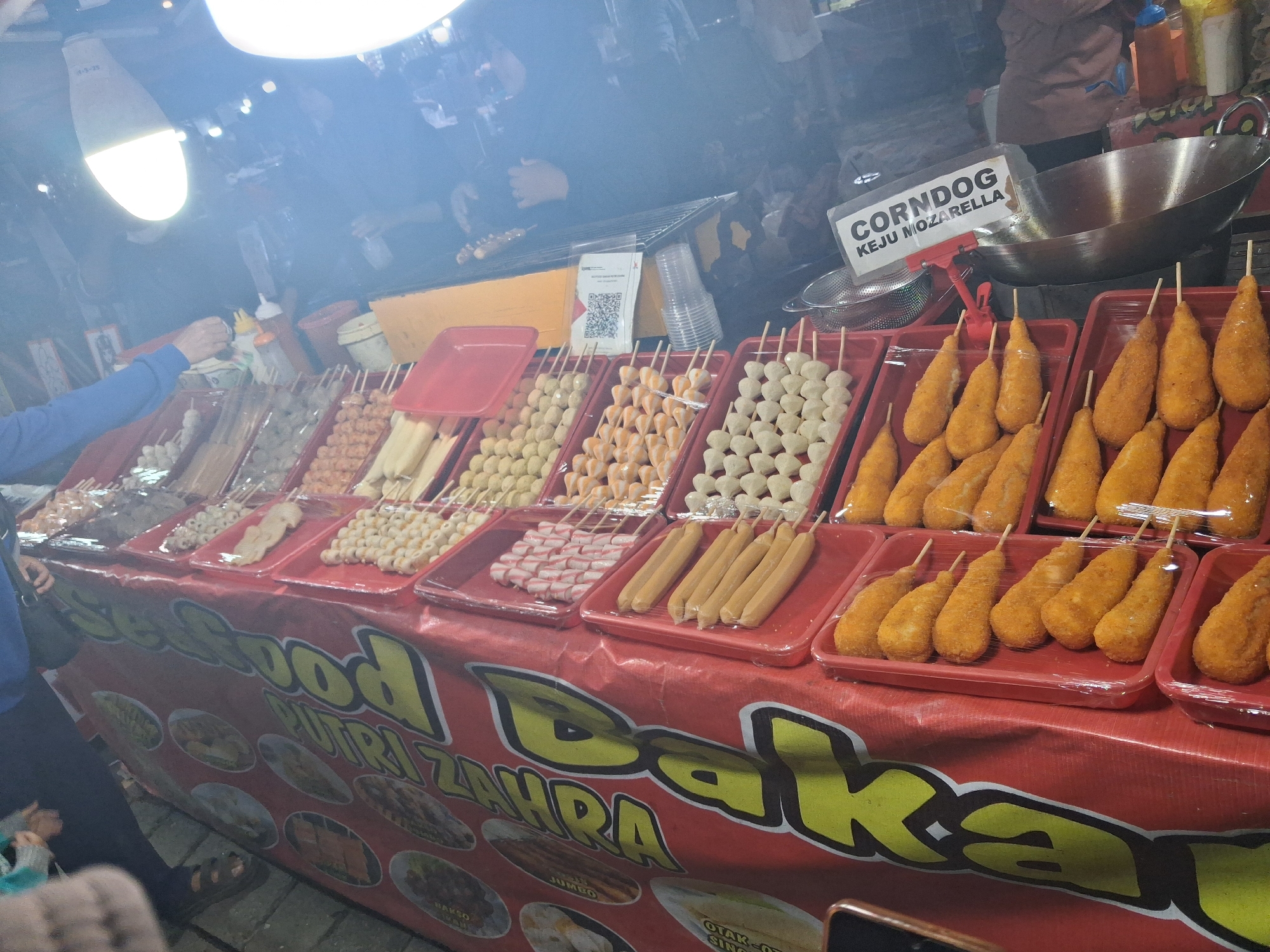 The image depicts a street food stall in Jambi, Indonesia, likely at a night market given the artificial lighting. The stall is adorned with a red banner featuring large yellow text that reads "SEAFOOD BAKAR ZAHRA," suggesting a specialization in grilled seafood. Various food items are displayed in red plastic trays, including what appear to be meatballs, fish cakes, sausages, and breaded corn dogs labeled "CORNDOG KEJU MOZARELLA." In the background, a large metal wok is visible, possibly used for frying. Several people are present, including a vendor or customer reaching for a bottle on the counter and another person partially visible in the foreground. The overall atmosphere is that of a bustling food vendor operation.