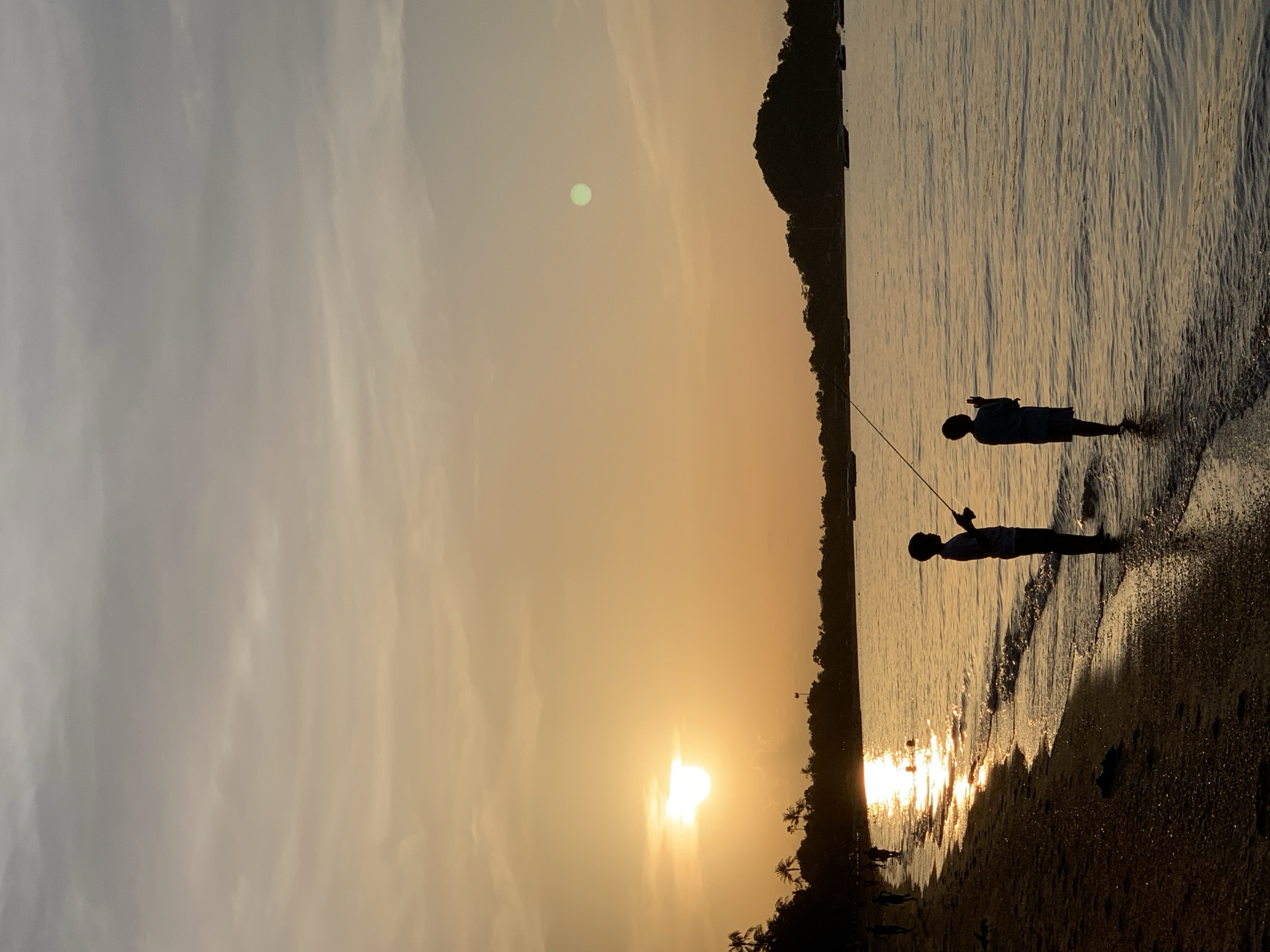 In the city of Ko Samui District, Thailand, during a golden hour sunset, two young boys are silhouetted against the water. One boy stands on the sandy shore, holding a fishing rod. The other boy stands closer to the water's edge, also holding a fishing rod, with gentle waves lapping at his feet. Palm trees and a dense treeline are visible in the background, silhouetted against the bright orange sky. Several boats are moored in the calm sea, and a small, dark island rises from the horizon. The sun, a bright orb, casts a shimmering reflection on the water. The sky transitions from a bright yellow near the horizon to a muted gray at the top of the frame.