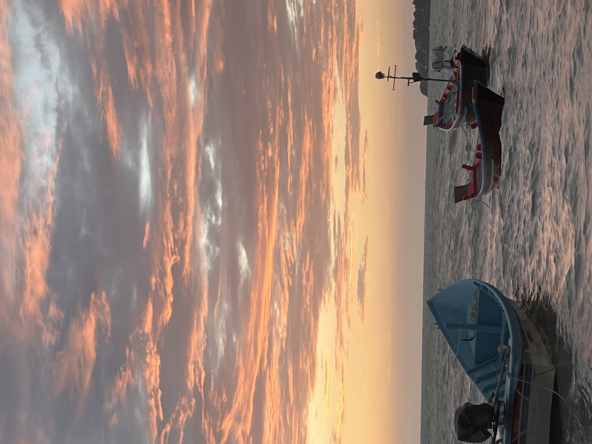 The image depicts a serene sunset scene at a beach in Ko Pha-ngan District, Thailand. The sky is a canvas of soft pink and orange hues, with scattered clouds creating a textured backdrop. Three boats are anchored in the gentle waves of the sea. In the foreground to the left, a light blue fishing boat with a black outboard motor is partially visible. To the right, two smaller boats, one red and blue and another with a dark hull and red trim, are also floating on the water. In the distance, a faint outline of a coastline with trees and buildings suggests the presence of land. The overall atmosphere is tranquil and picturesque, characteristic of a peaceful evening by the sea.