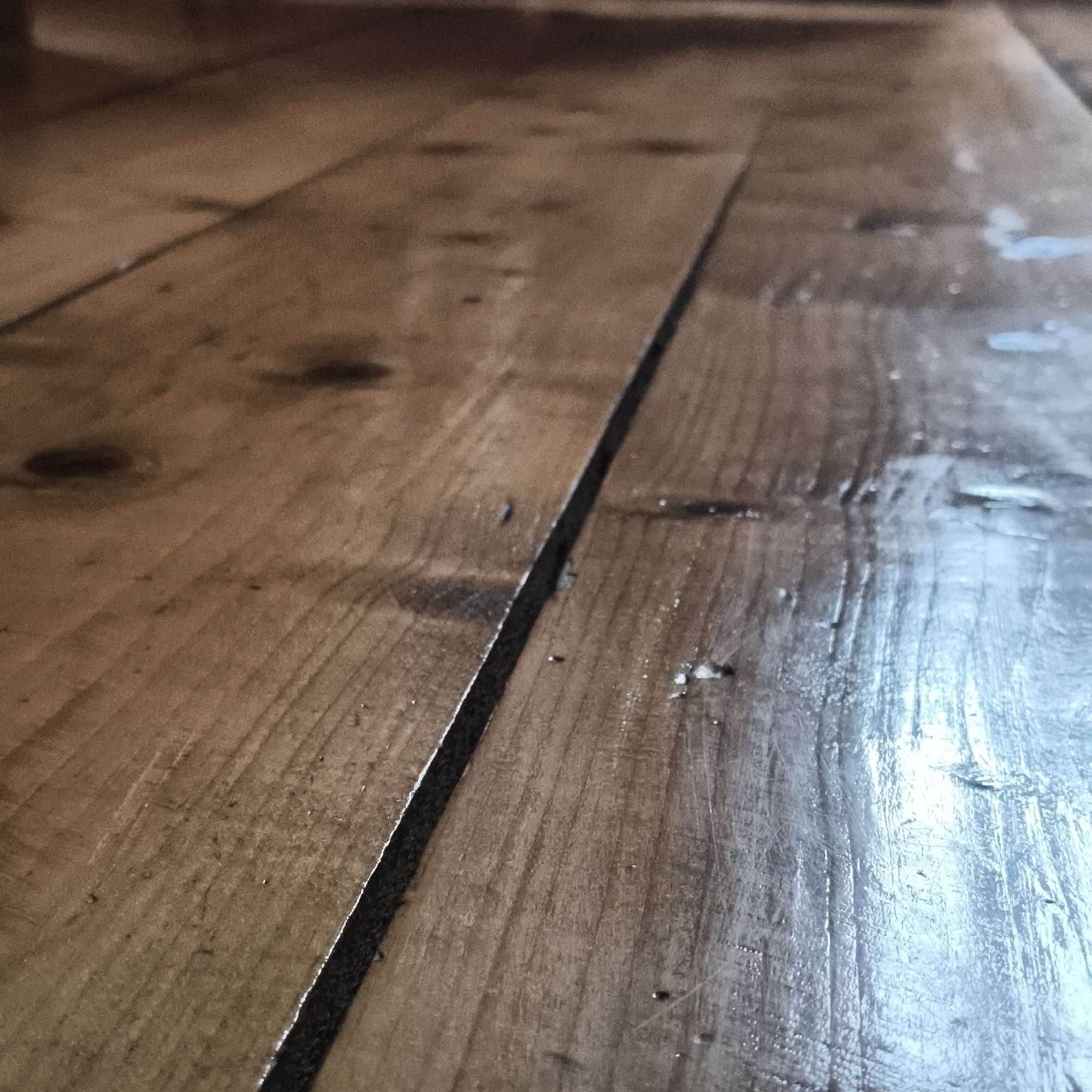 The image shows a close-up of a wooden floor with noticeable gaps between the planks. The wood has a warm, brown tone, and some areas appear shiny or wet, possibly due to a recent cleaning or sealant application. There are visible knots and imperfections in the wood grain. The perspective is angled, creating a sense of depth. No specific elements in the image suggest a direct connection to Chaikovsky, Russia.