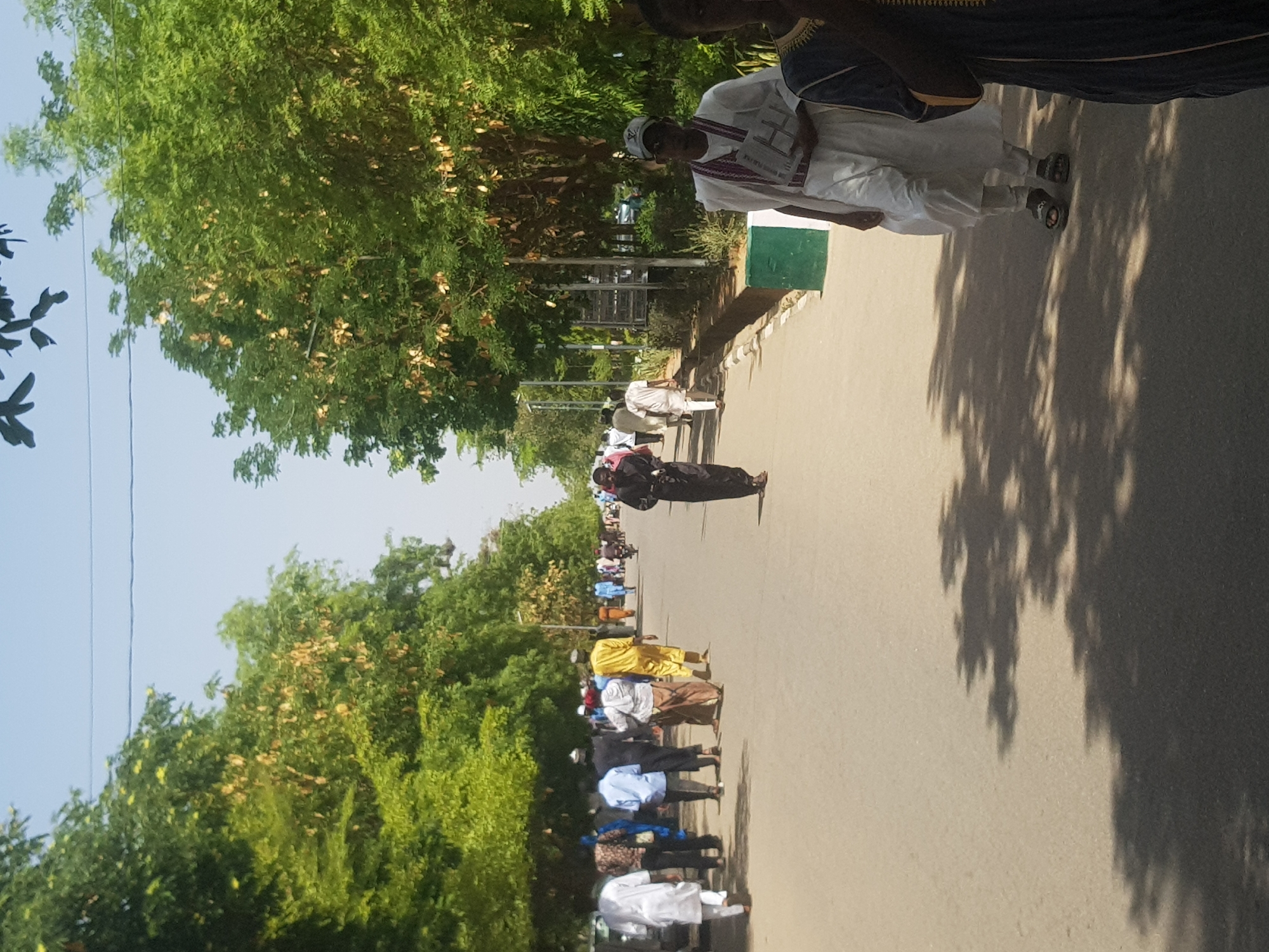 A medium, eye-level shot shows a tree-lined street in Dabawa, Nigeria, on a bright, sunny day, where a procession of people are standing in the road. The street extends into the distance, lined with lush green trees on both sides, creating a canopy. The light blue sky is visible between the tree branches.
A line of people stands along the right side of the street, dressed in various traditional or religious attires. They include a man in a white robe with a red scarf, another in black, and others in colorful shirts. The people appear to be lined up for a religious or cultural event.
The scene includes a green and white post on the side of the road. The road is paved and stretches into the distance. The image is slightly rotated.