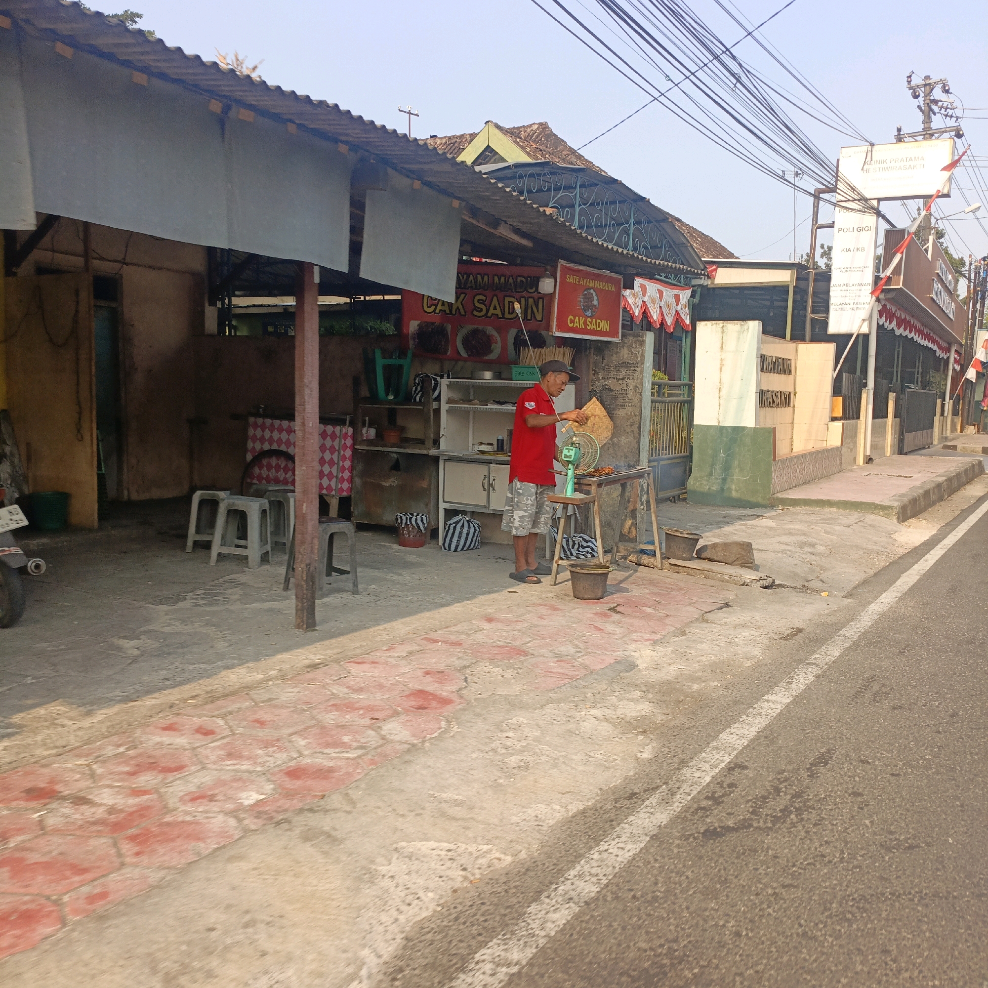 The image captures a street-level view of a small food stall in Salatiga, Indonesia. The stall, identified by signage as "CAK SADIN," appears to specialize in "SATE AYAM MADURA" (Madurese chicken satay). A man wearing a red shirt and patterned shorts is actively preparing food, fanning a grill with a woven fan. In the foreground, a paved walkway with red hexagonal tiles runs alongside the road. The stall itself is simple, with a corrugated metal roof supported by wooden beams. Behind the stall, a row of white stools is visible. To the right of the food stall, a sign for "KLINIK PRATAMA HE STI WIRASAKTI" indicates the presence of a clinic. The environment is characterized by an overcast sky and a mix of residential and commercial buildings lining the street. A motorcycle is parked on the left side of the image. The overall scene depicts a typical urban street scene in Indonesia, with an emphasis on local commerce and daily life.