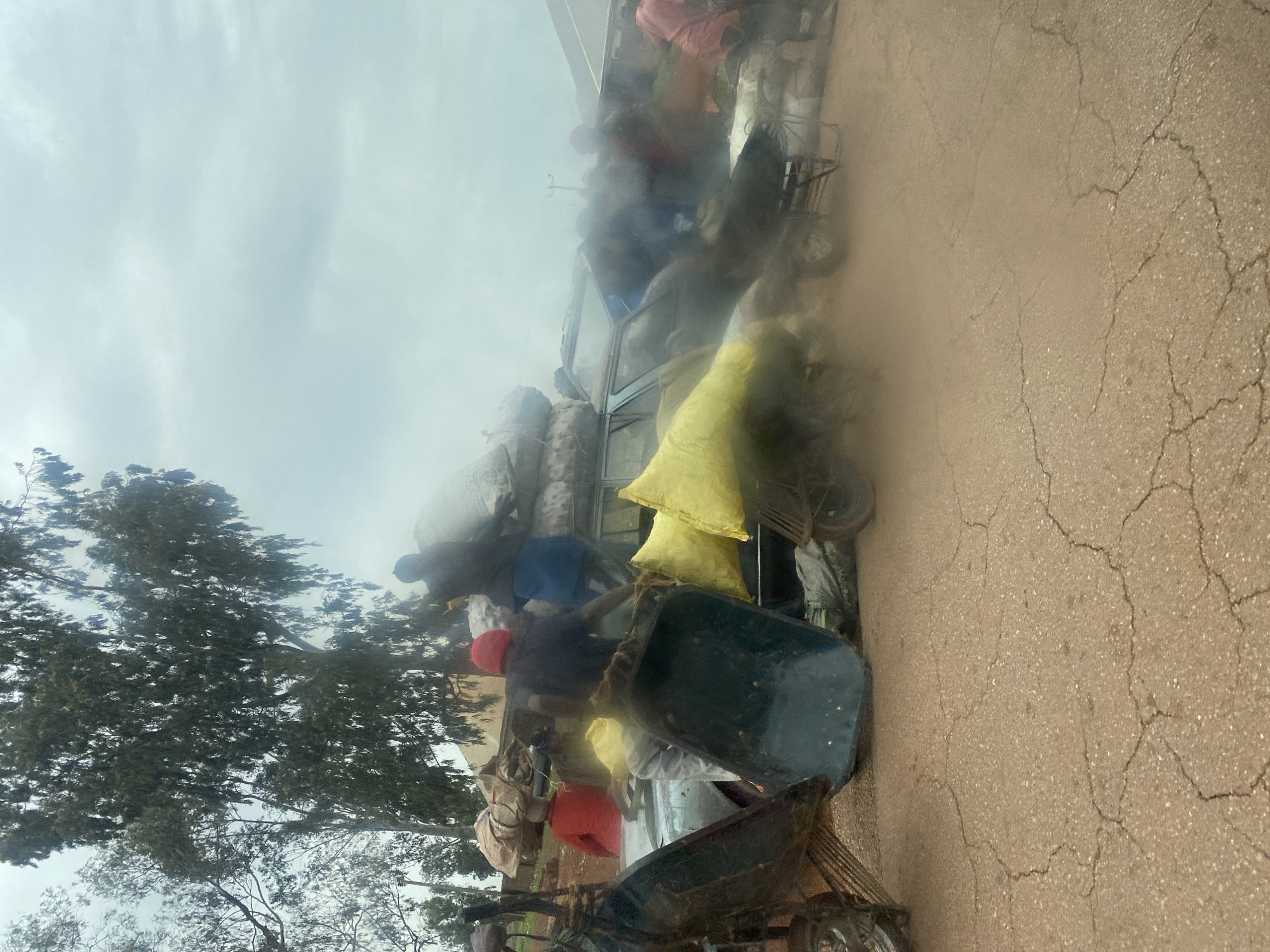 The image shows a scene in what appears to be Durimi, Nigeria, featuring a heavily loaded van or small truck. The vehicle is piled high with large sacks or bags, possibly containing goods or cargo. Several individuals are present around the vehicle, seemingly involved in the loading or unloading process. Some are on top of the load, while others are on the ground near the truck and moving carts. The setting includes a cracked pavement road and trees on the side, suggesting a rural or semi-rural environment. The sky is overcast, indicating potentially poor weather conditions.
