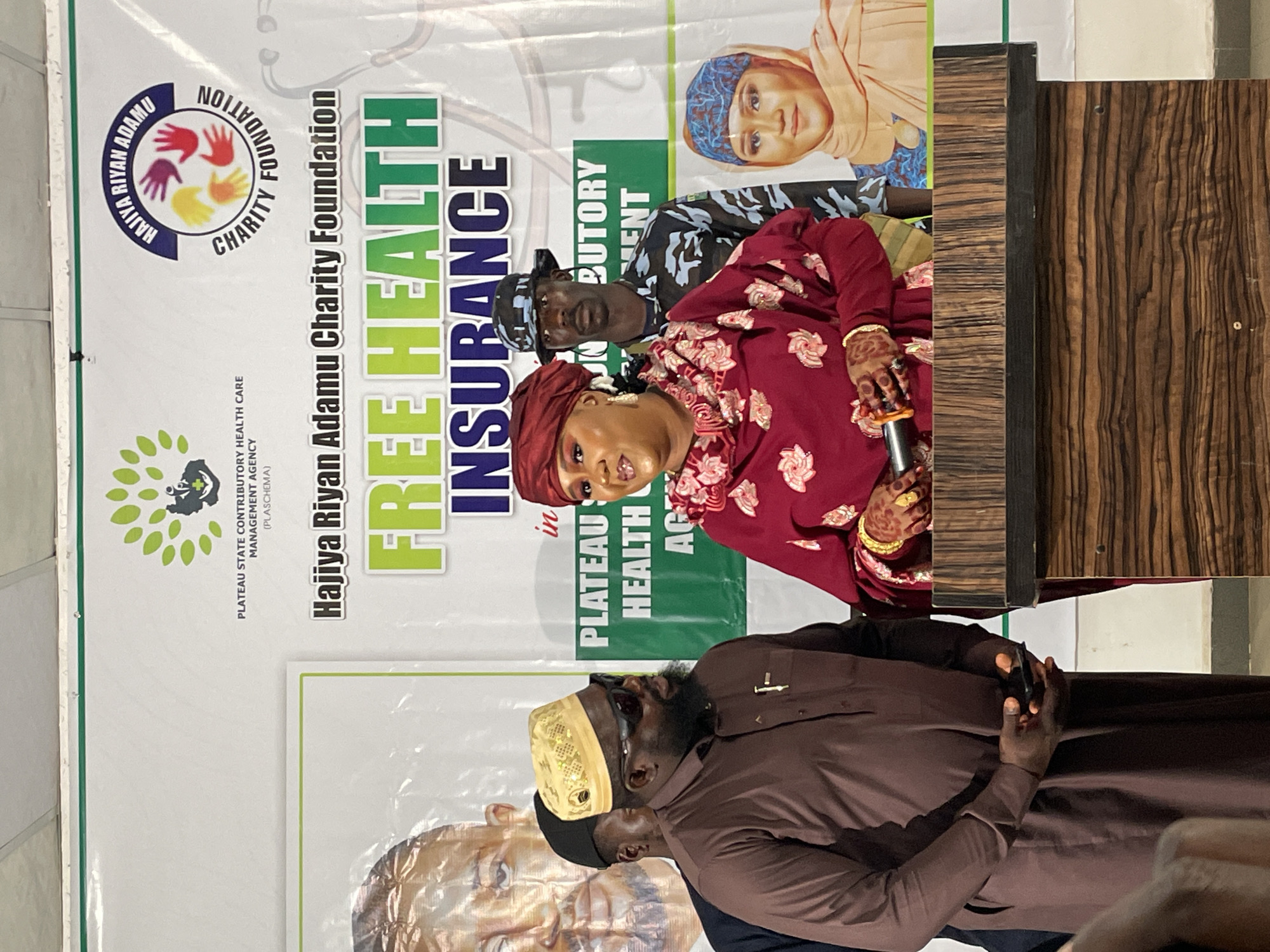 The image captures a scene at what appears to be a public health-related event. A backdrop displays the logos of the "Hajiya Riyan Adamu Charity Foundation" and "Plateau State Contributory Health Care Management Agency (PLASCHEMA)," alongside the words "FREE HEALTH INSURANCE". Three people are visible in the foreground. A woman is seated at a podium, holding a microphone and speaking. A man in uniform is behind her. Another man, wearing traditional attire, is standing to the side. The image suggests an event in Jos, Nigeria, focused on free health insurance, possibly organized by the charity in collaboration with the Plateau State health agency.