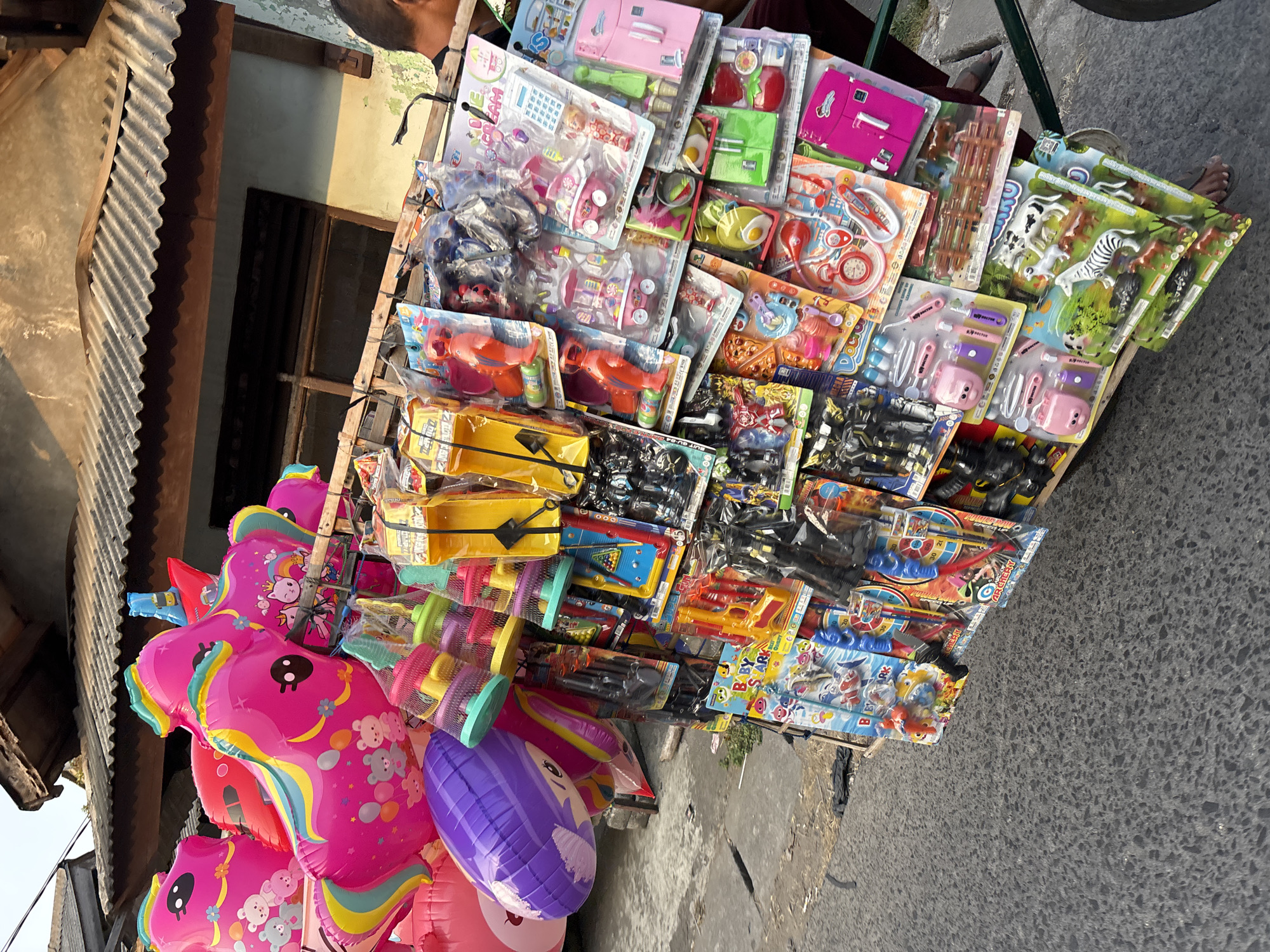 In Bekasi, Indonesia, a street vendor displays a colorful assortment of toys on a cart. The cart is laden with various items, including bright pink unicorn balloons, purple balloon characters, and numerous packaged toys. These packaged toys include what appear to be toy doctor kits, ice cream carts, refrigerators, toy guns, animal figures, and other assorted plastic playthings. The cart is positioned outdoors on a paved surface, likely a sidewalk or street, with a corrugated metal roof structure visible in the background, suggesting a building or stall. A person's legs and feet are visible on the right side of the image, indicating their presence as the vendor or a passerby. The overall scene depicts a typical street market environment where small toys are sold.