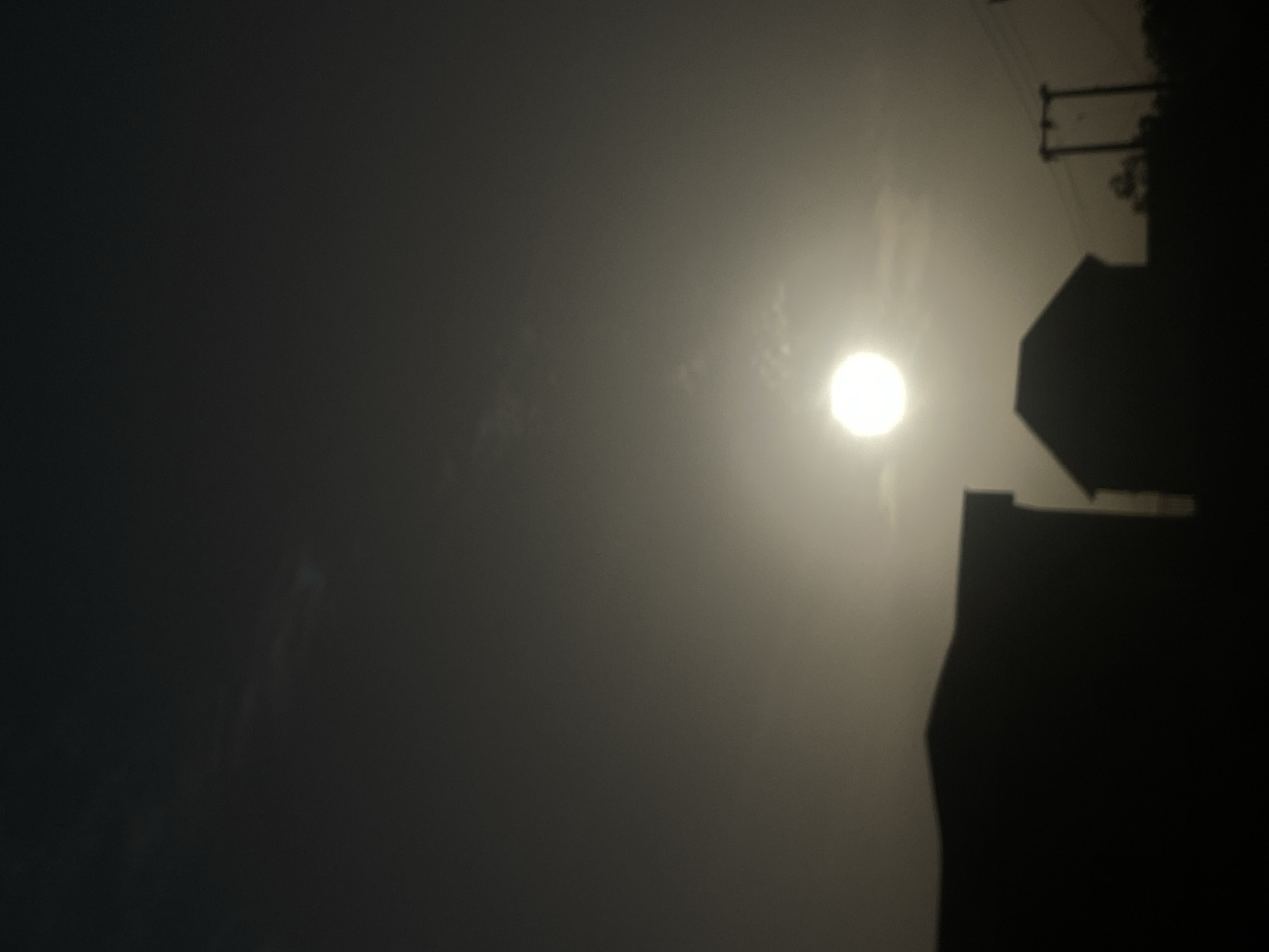 The image, taken in Kwarbai B, Nigeria, captures a hazy outdoor scene dominated by a brilliant, diffused sun. The sun appears as a bright, circular disc, positioned towards the upper right, suggesting either early morning or late afternoon light. The sky around the sun is enveloped in a soft, bright haze, indicating foggy, misty, or dusty atmospheric conditions that diffuse the light and obscure clear details.
On the right side of the frame, dark silhouettes of man-made structures are prominently visible. A distinct, angular outline suggests the roofline of a building, rendered in stark shadow against the bright sky. Above and behind this structure, the dark shapes of utility poles or power line towers, complete with horizontal crossbars and faint connecting lines, are also silhouetted. Some indistinct, dark forms, likely trees or foliage, are integrated with these structures.
The rest of the sky transitions from the bright, hazy glow near the sun to much darker tones, ranging from deep gray to almost black, particularly on the left side of the image. The strong backlighting creates a high-contrast composition, making all elements other than the sun appear as undifferentiated silhouettes. There is no discernible human or animal activity, and no visible text in the image. The scene evokes a quiet, atmospheric moment under low-visibility conditions.