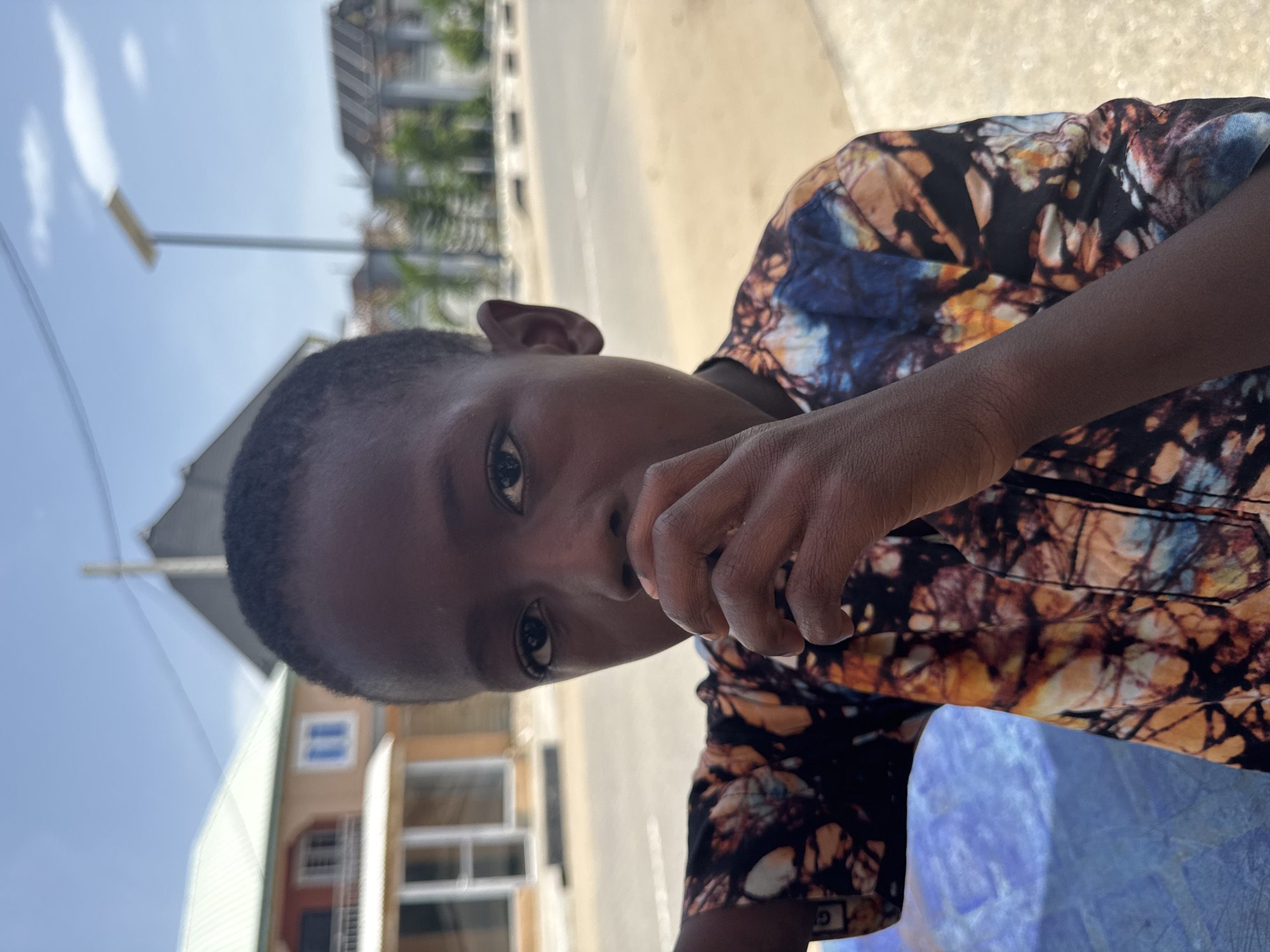 A person with dark skin and short, dark hair is depicted in a close-up shot. The person is wearing a multi-colored shirt with a tie-dye pattern in shades of black, blue, and brown. Their hand is raised towards their mouth. In the background, there is a street, buildings, and a blue sky. The location is Badagry, Nigeria.