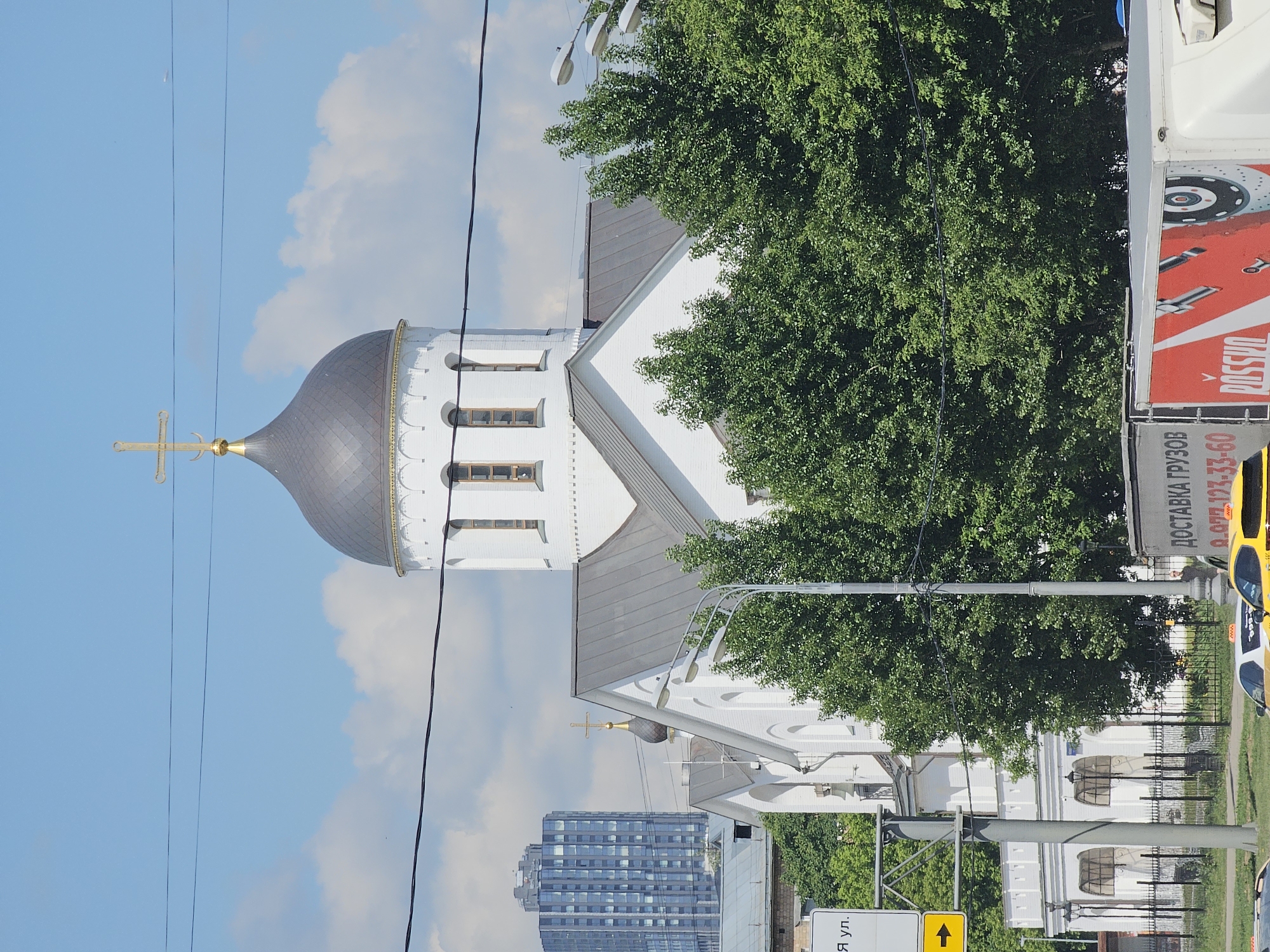 The image displays an urban scene featuring a white Orthodox church under a bright blue sky with scattered white clouds. The church has a prominent dark, possibly grey, dome crowned with a golden cross. Its architecture includes a tiered bell tower with arched and rectangular windows. Lush green trees partially obscure the church, dominating the right side of the frame and appearing in the foreground.
In the background, to the left of the church, a modern high-rise building with numerous windows is visible, contrasting with the traditional religious architecture. Several power lines stretch across the sky, adding to the urban feel.
On the far right, a section of a red and white truck or trailer is visible, featuring Cyrillic text advertising "ДОСТАВКА ГРУЗОВ" (Delivery of goods), a phone number "8 (927) 93-60", and "РОССИЯ" (Russia), along with a graphic of a brake disc. Below this truck, the roof of a yellow taxi car can be seen. At the bottom of the image, a yellow road sign with a black arrow pointing forward is partially visible, displaying the Cyrillic letters "Я УЛ." (likely part of a street name, e.g., "-ya Street"). Parts of streetlights and a black metal fence are also present. The scene is set during the daytime, under sunny weather, depicting a blend of historic and modern elements typical of a city like Moscow.