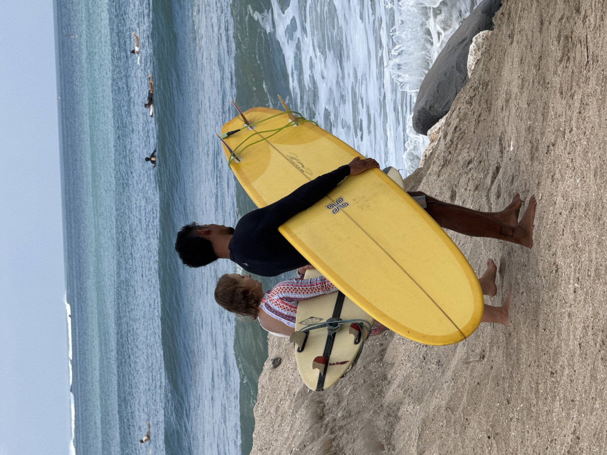 Two people, a man and a woman, are walking on a beach with surfboards. The man is on the right, carrying a large yellow surfboard. He is wearing a black long-sleeved shirt. The woman is on the left, carrying a smaller, lighter-colored surfboard. She is wearing a colorful patterned dress. The beach has sand and rocks. In the background, there are ocean waves and several other surfers in the water. The scene appears to be in Kuta, Indonesia, a popular surfing destination known for its beaches and waves. The sky is overcast, and the water is a mix of blue and green.