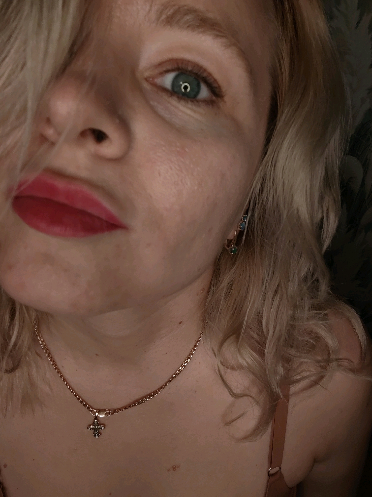 The image is a close-up portrait of a woman's face and upper body. She has blonde hair that is partially covering her face, and she is wearing red lipstick and a necklace with a cross pendant. There are also earrings visible on her left ear. The background is dark and appears to be a room. There is no information in the image to suggest a location in Izhevsk, Russia.