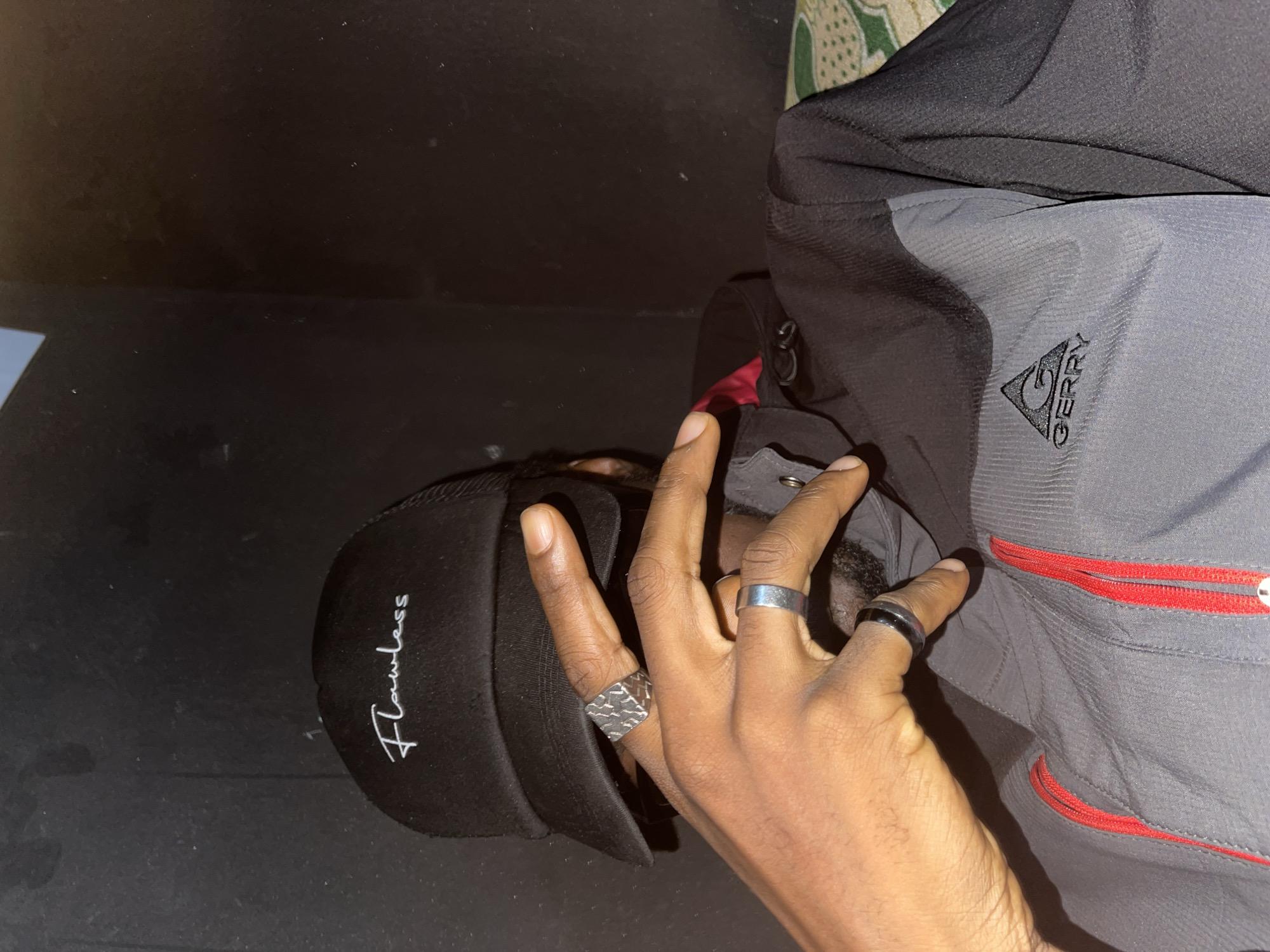 A medium shot image shows a person from the shoulders up. They are wearing a black baseball cap with white cursive writing that says "Flawless". The person has dark skin and is wearing rings on their fingers. They are also wearing a grey and black jacket with red zippers, and a Gerry logo on the left side of the chest. The background is dark and appears to be a wall. The image does not provide enough information to determine the location in the city of Jos, Nigeria.