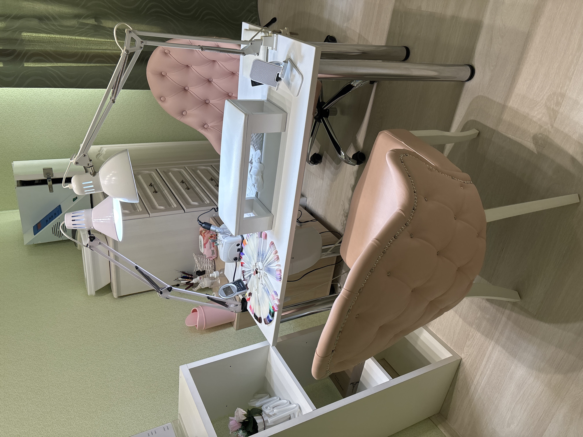 The image shows a nail salon setup. Two manicure tables with white tops and integrated storage are present. One table has a built-in drawer with cotton balls, and both have adjustable lamps. The chairs are upholstered in a tufted pink material. Various manicure tools and products are visible on the tables. The floor is light green with a subtle pattern. The location is Kostoryata, Russia.