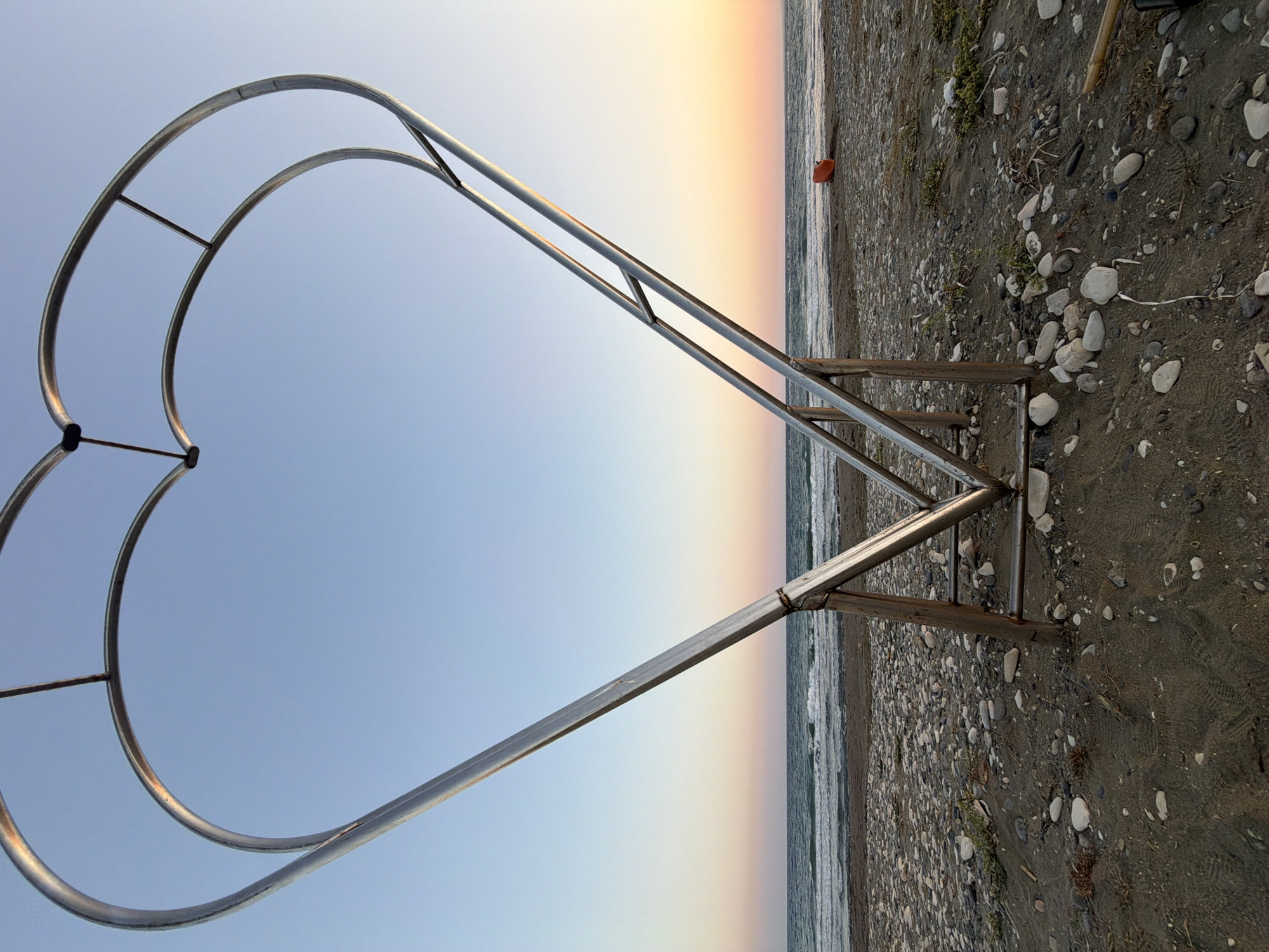 A large, metal heart-shaped sculpture is positioned on a pebble and sand beach in Meneou, Cyprus. The sculpture's frame, made of silver-colored metal, is open and forms the outline of a heart. It is supported by wooden posts and metal bracing. The beach is covered in dark sand and smooth, light-colored pebbles. In the background, the sea meets the horizon, with gentle waves rolling onto the shore. The sky above is a clear, light blue, transitioning to soft hues of pink and orange near the horizon, indicating either sunrise or sunset. A small, bright orange object, possibly a buouy or marker, is visible on the beach to the right of the sculpture. The overall scene is tranquil and scenic.