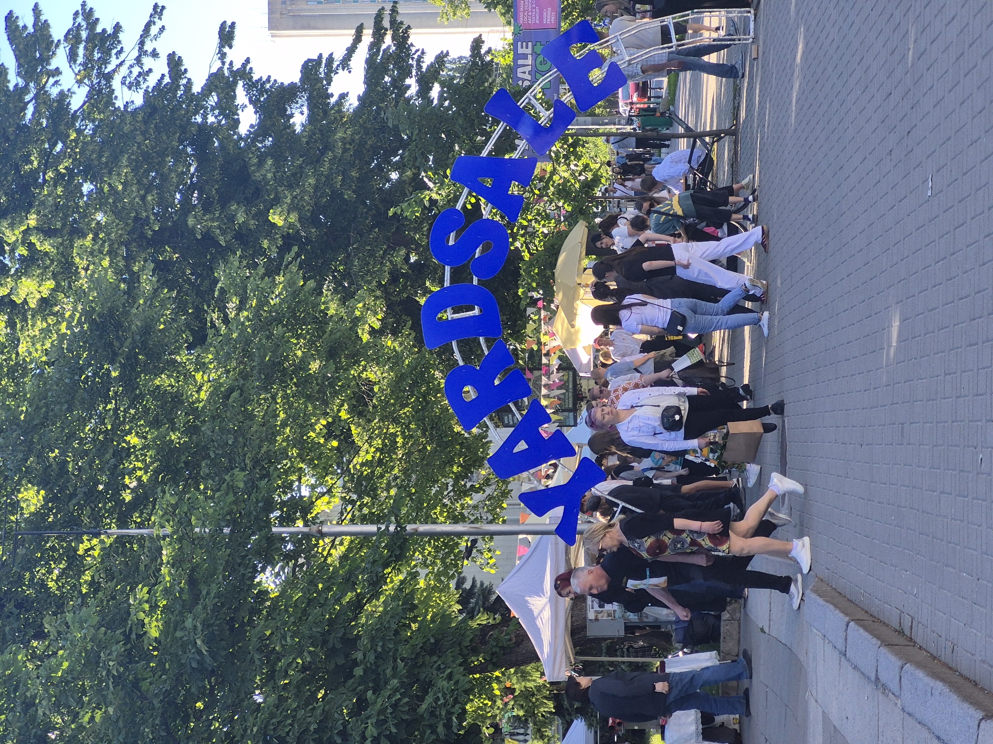 The image depicts an outdoor yard sale event taking place in Chișinău, Moldova, under a large, arching blue sign that reads "YARDSALE". The scene is bustling with people of various ages browsing through stalls set up under white tents. The ground is paved with gray cobblestones, and the event is shaded by large, mature trees with lush green foliage. In the background, a tall, modern building is visible, suggesting an urban setting. The atmosphere appears to be lively and social, with individuals engaging in shopping and conversation.