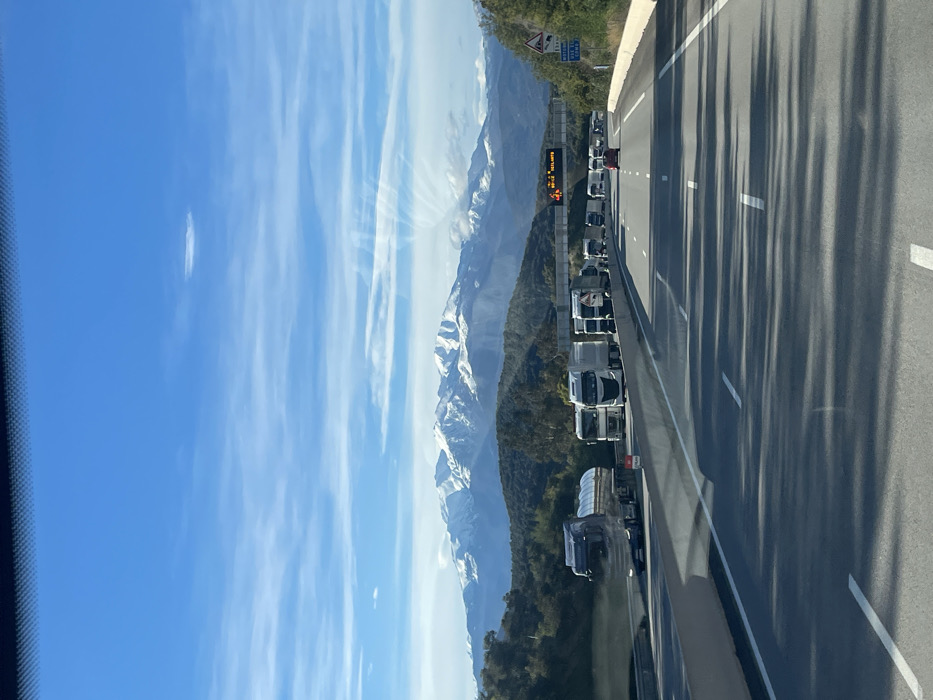 The image presents a daytime view from inside a vehicle on a multi-lane highway, likely the A9 autoroute near Le Perthus, France, facing generally south towards the Pyrenees mountains.
The sky is predominantly clear blue with scattered wispy white clouds and a distinct, elongated lenticular cloud centered in the upper frame. In the background, a majestic range of snow-capped mountains, consistent with the Pyrenees, stretches across the horizon, flanked by forested hillsides.
The highway features at least three lanes in the direction of travel, separated by a concrete median barrier. The leftmost lane appears relatively clear. The middle and right lanes exhibit significant traffic congestion, primarily composed of commercial semi-trucks. Visible trucks include a silver tanker truck in the left-center of the foreground and numerous white tractor-trailer units extending into the distance. A red passenger vehicle is discernible amidst the truck traffic further ahead in the middle lane.
Overhead, a gantry sign spans the highway, displaying amber text and an orange warning icon, likely indicating traffic congestion or advisories. On the right shoulder, a blue directional sign shows "10 km" and an arrow pointing right, with smaller text below, possibly indicating a route or destination. A triangular warning sign is positioned beneath it. A small red sign is visible on the median barrier in the foreground, possibly indicating a temporary stop or warning. Bright sunlight casts long shadows of vehicles and roadside structures across the asphalt pavement. No individuals are clearly visible.