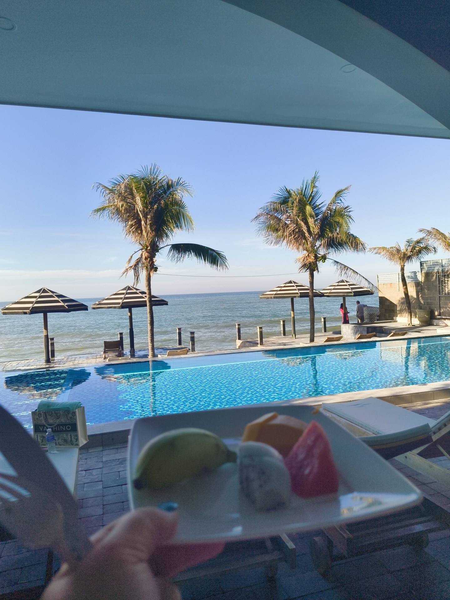 The image depicts a resort setting with a swimming pool and the ocean in the background. In the foreground, a plate holds a selection of fruits, including what appears to be dragon fruit, mango, and watermelon, held by a person's hand with blue nail polish. A fork and knife are also visible. The pool, with its mosaic tiles, reflects the bright blue sky and surrounding palm trees. Two striped umbrellas stand near the pool, casting shadows. Palm trees are scattered throughout the scene, with some taller ones in the background. The ocean stretches to the horizon under a clear blue sky. Two individuals, one wearing a red garment and the other a grey shirt, are visible near the umbrellas in the middle ground. A table with a shopping bag labeled "VAHINO" and a bottle of water is positioned to the left of the fruit plate. The scene is set on a sunny day. The location is identified as Phu Thuy Ward, Vietnam.