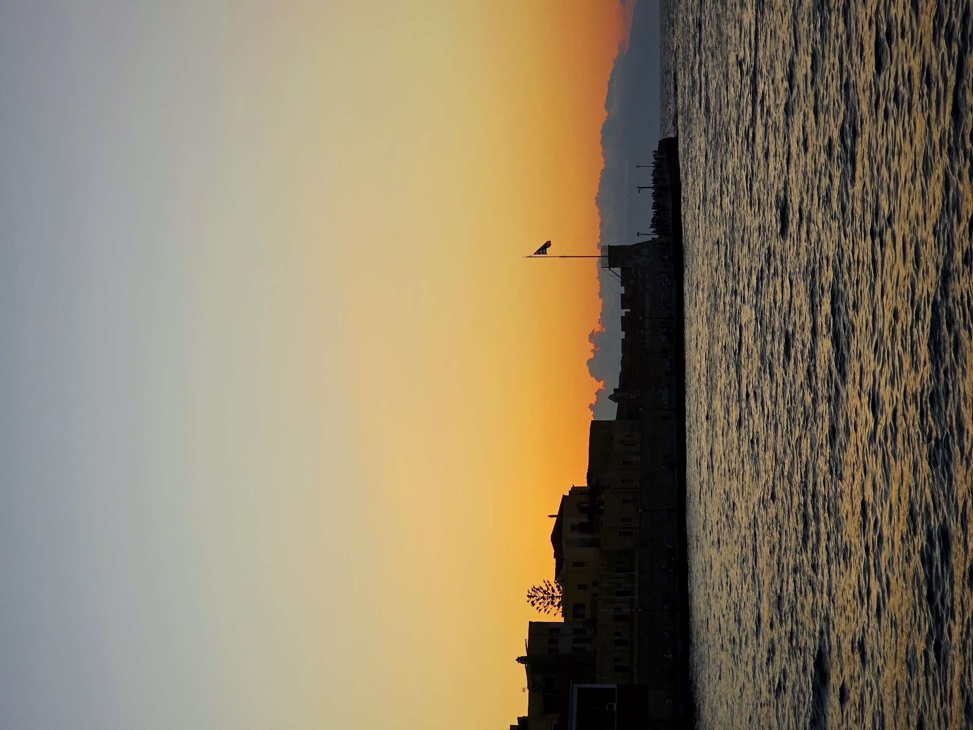 At sunset, a silhouetted coastal fortification, likely part of the Venetian Harbor in Chania, Greece, stands against a gradient sky of soft blues and vibrant oranges. The water in the foreground reflects the warm hues of the sky, creating a textured, dark surface with glints of light. Along the top of the fortification, a line of figures, likely tourists or locals, are visible, observing the view. A flagpole with a flag, possibly the Greek flag, stands prominently on a tower-like structure. The buildings behind the wall are also silhouetted, with a distinctive tree shape adding a natural element to the man-made structures. The overall scene captures a serene evening ambiance in a historic seaside location.