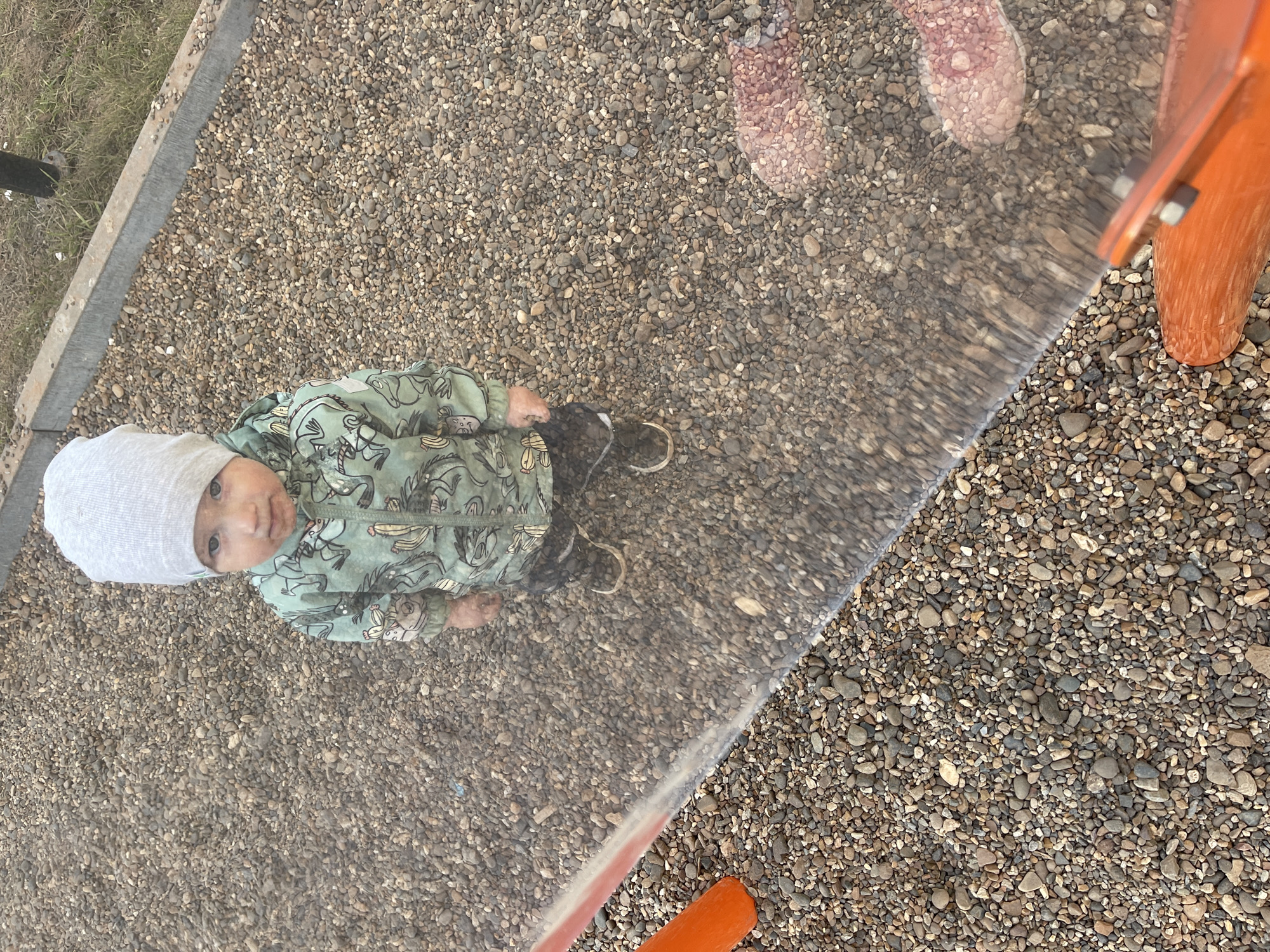 A child wearing a grey hat and a green jacket with animal designs stands on a surface covered in small pebbles. The child is looking up. Part of an orange structure is visible on the right side of the image. A transparent barrier, perhaps glass or plastic, runs diagonally across the scene, partially obscuring the pebbles and the child. There are footprints on the upper right behind the glass. The location is likely a playground, given the pebbled ground and the orange structure. Without further information, confirming the location as Izhevsk, Russia, is impossible.
