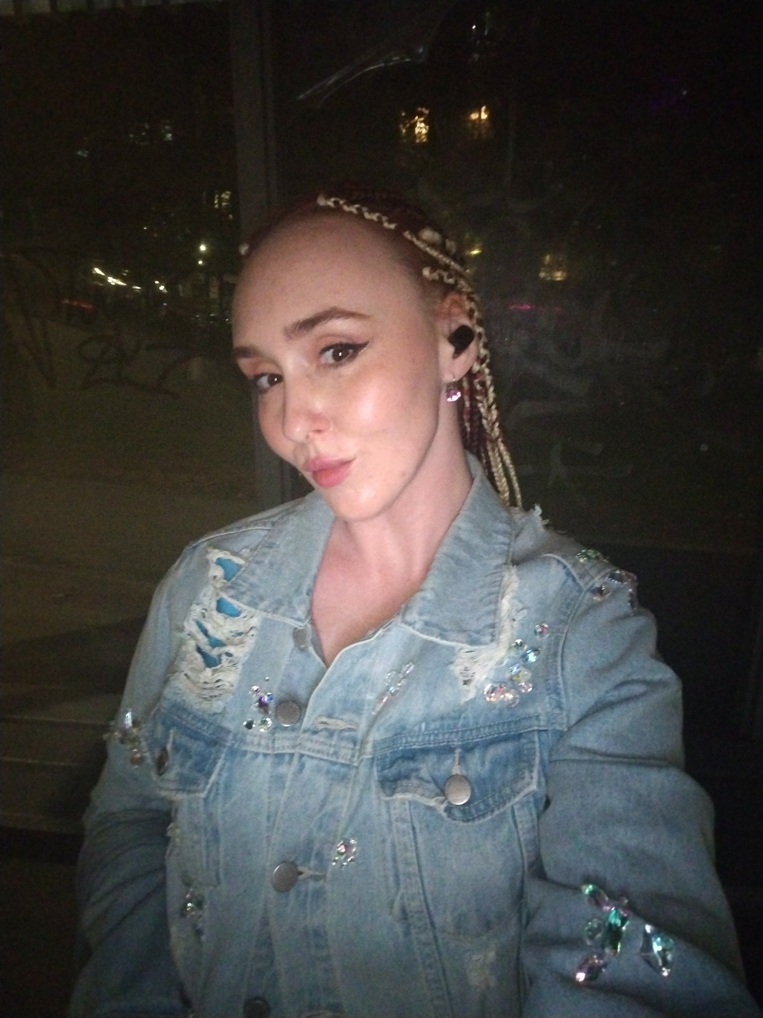 A person is standing indoors, likely near a window, based on the city lights visible through the glass behind them. The location is described as Izhevsk, Russia. The person appears to be female with light skin, light-colored hair styled in braids, and is wearing a denim jacket. The jacket is adorned with decorative embellishments, including what appear to be rhinestones. The person is wearing an earbud in one ear and has visible earrings. The image is relatively dark.