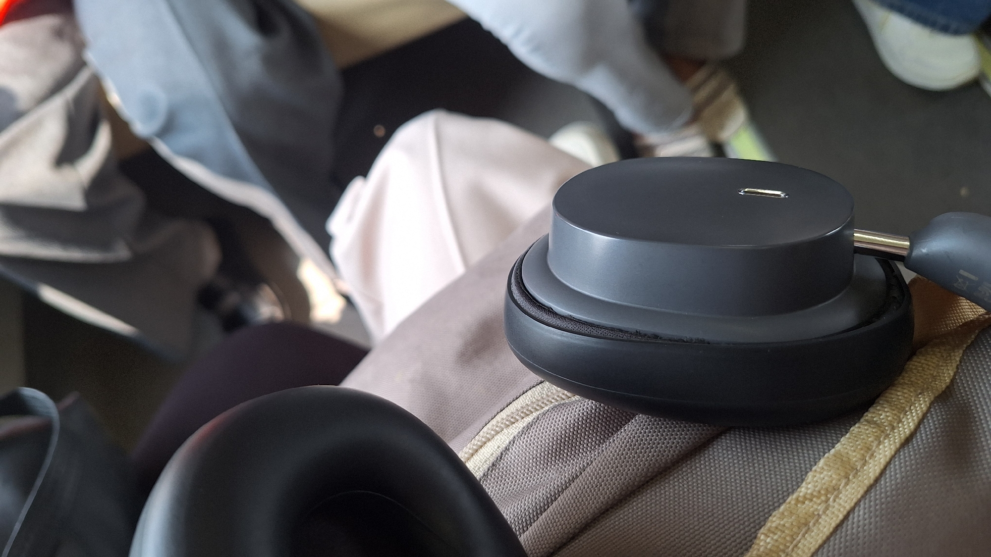 A close-up shot shows a pair of black wireless headphones resting on a textured grey fabric, likely a bag or clothing. The headphones are partially visible, with the ear cup and a portion of the headband in focus. The top of the right ear cup features a metallic port. The background consists of out-of-focus items including clothing and bags. The location is implied to be Karangsari, Indonesia.