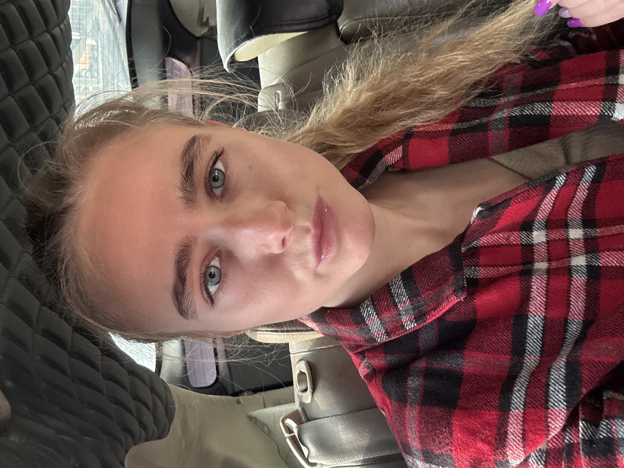 A portrait-style image captures a young woman from the chest up, positioned at a slight angle. She has fair skin, light-colored hair that appears blonde or light brown, styled loosely, and striking blue-grey eyes that look directly at the viewer. Her eyebrows are well-defined, and her lips have a natural, glossy appearance, slightly parted. She has a neutral to slightly contemplative expression.
She is wearing a red, black, and white plaid shirt, visible on her torso and sleeve. One of her fingernails, visible on the right side of the frame near her shoulder, is painted a vibrant purple.
The setting appears to be the interior of a vehicle, likely a car. To her left, a black, quilted pattern, possibly part of a car seat or headrest, is visible. Behind her, and to the right, lighter-colored upholstery, possibly beige or tan, and a metallic seatbelt buckle are discernible. The background is somewhat blurred, but suggests an indoor or enclosed space with some ambient light.
The image seems to be a selfie or a close-up photograph taken while she is seated. No text is visible in the image.