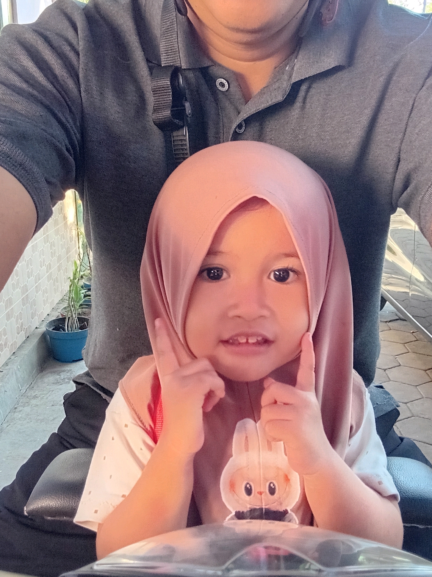 In Gebang, Indonesia, a young child wearing a pink hijab and a shirt with a rabbit appliqué is seated on a vehicle. The child is looking at the camera and has their fingers near their cheeks. Behind the child, an adult wearing a grey polo shirt is visible. The background shows a tiled wall and a potted plant. The ground is paved with hexagonal tiles. The scene appears to be outdoors, possibly on a street or pathway.