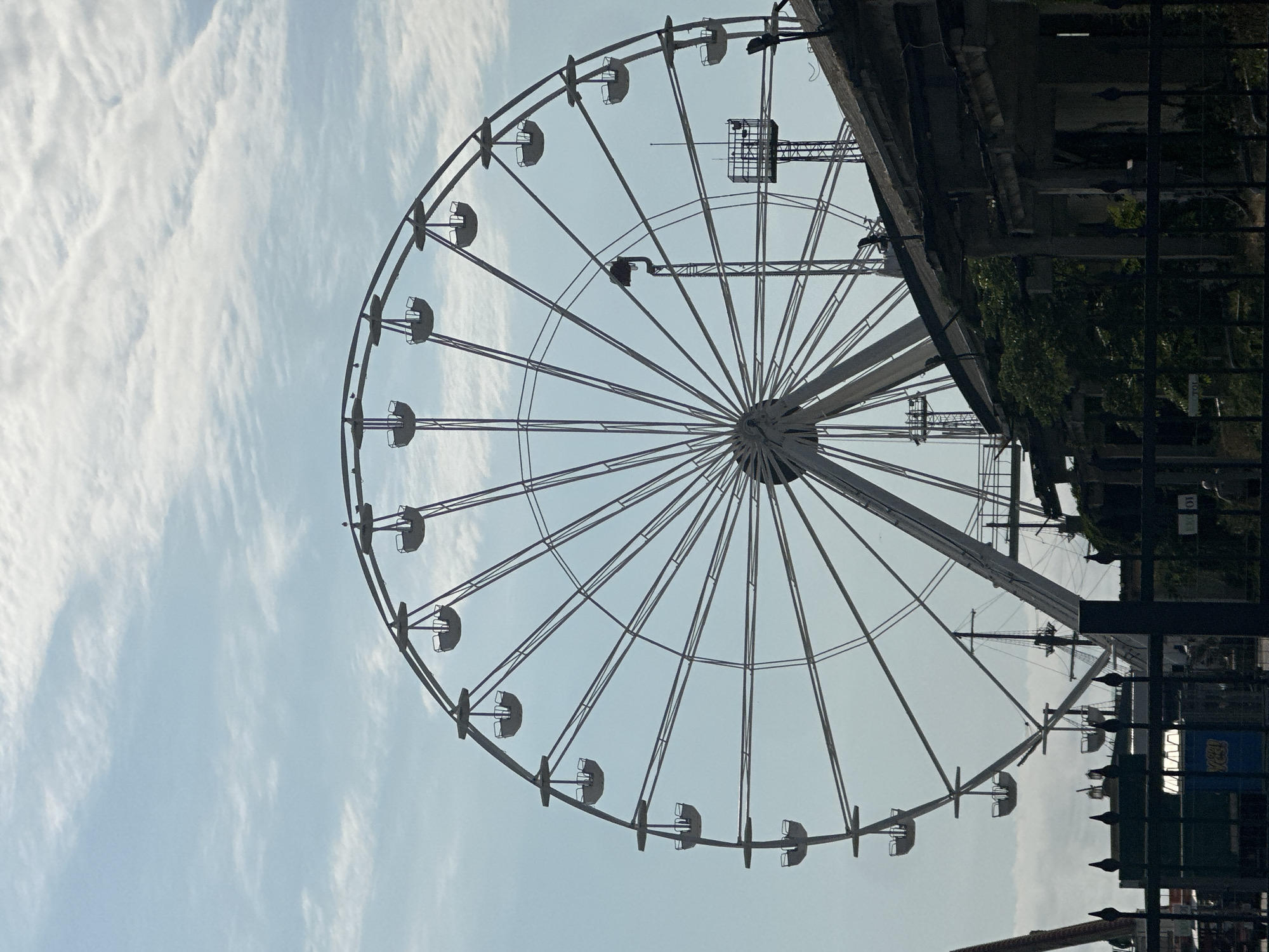 A Ferris wheel dominates the frame, silhouetted against a hazy blue sky with scattered wispy clouds. The Ferris wheel is large and intricate, with numerous spokes radiating from the center and many gondolas hanging from the outer rim. In the foreground, the roofline of a dilapidated building runs diagonally across the lower right portion of the image, obscuring the base of the Ferris wheel. The building appears to be made of weathered wood and has a dark, aged appearance. Behind the Ferris wheel and to the left, the masts and rigging of a tall ship are visible, suggesting a waterfront or harbor location. A black metal fence runs along the very bottom of the image, partially obscuring a low wall and some greenery. The overall impression is one of a slightly neglected or aged amusement park or seaside attraction.
