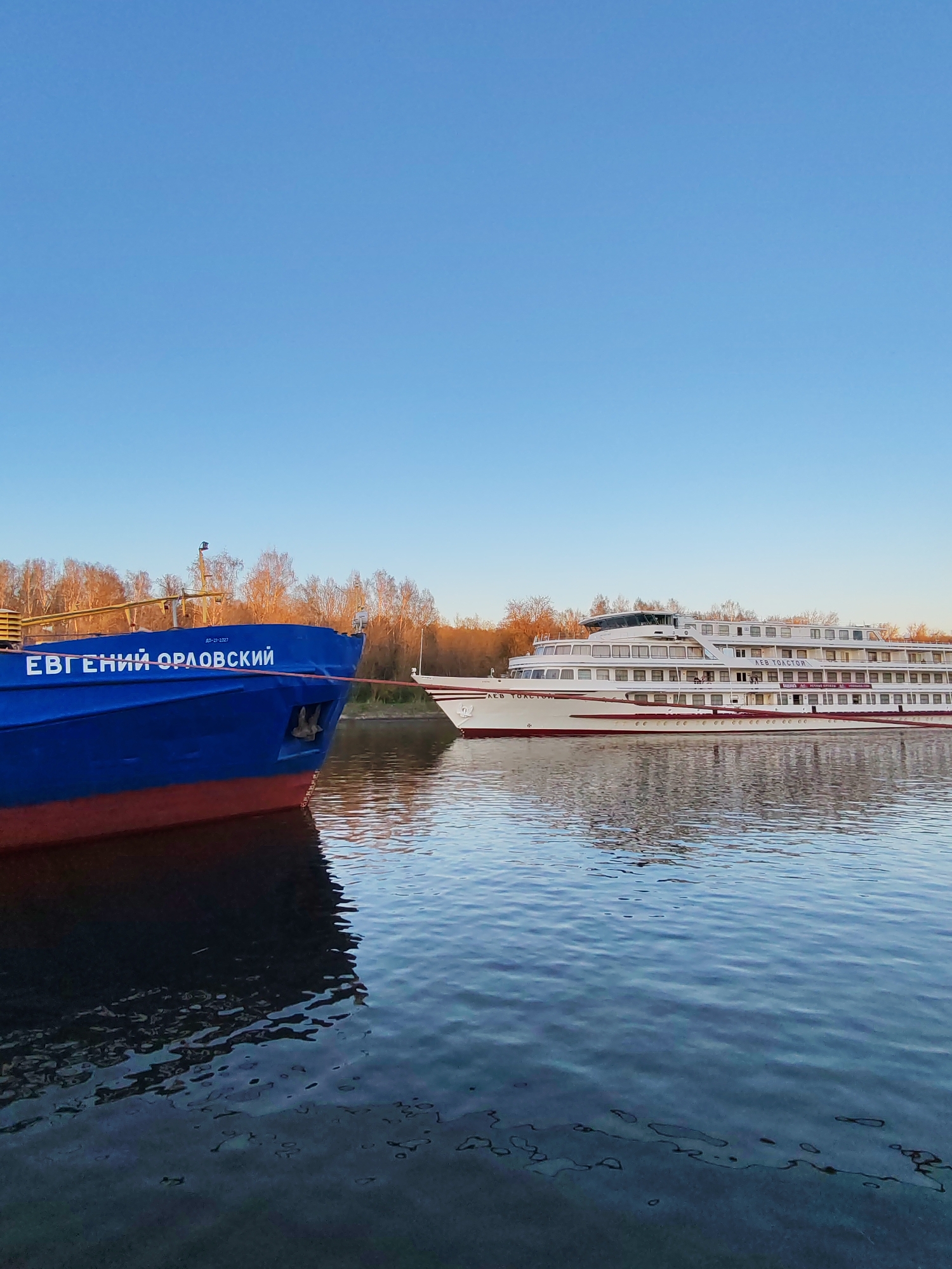 The image shows two boats on a body of water in Yakhroma, Russia. The first boat, on the left, is blue with a red waterline and has the name "ЕВГЕНИЙ ОРЛОВСКИЙ" written on its side. The second boat, on the right, is white with a red stripe and appears to be a larger passenger vessel. The water is calm with slight ripples. The background features a line of trees under a clear blue sky.
