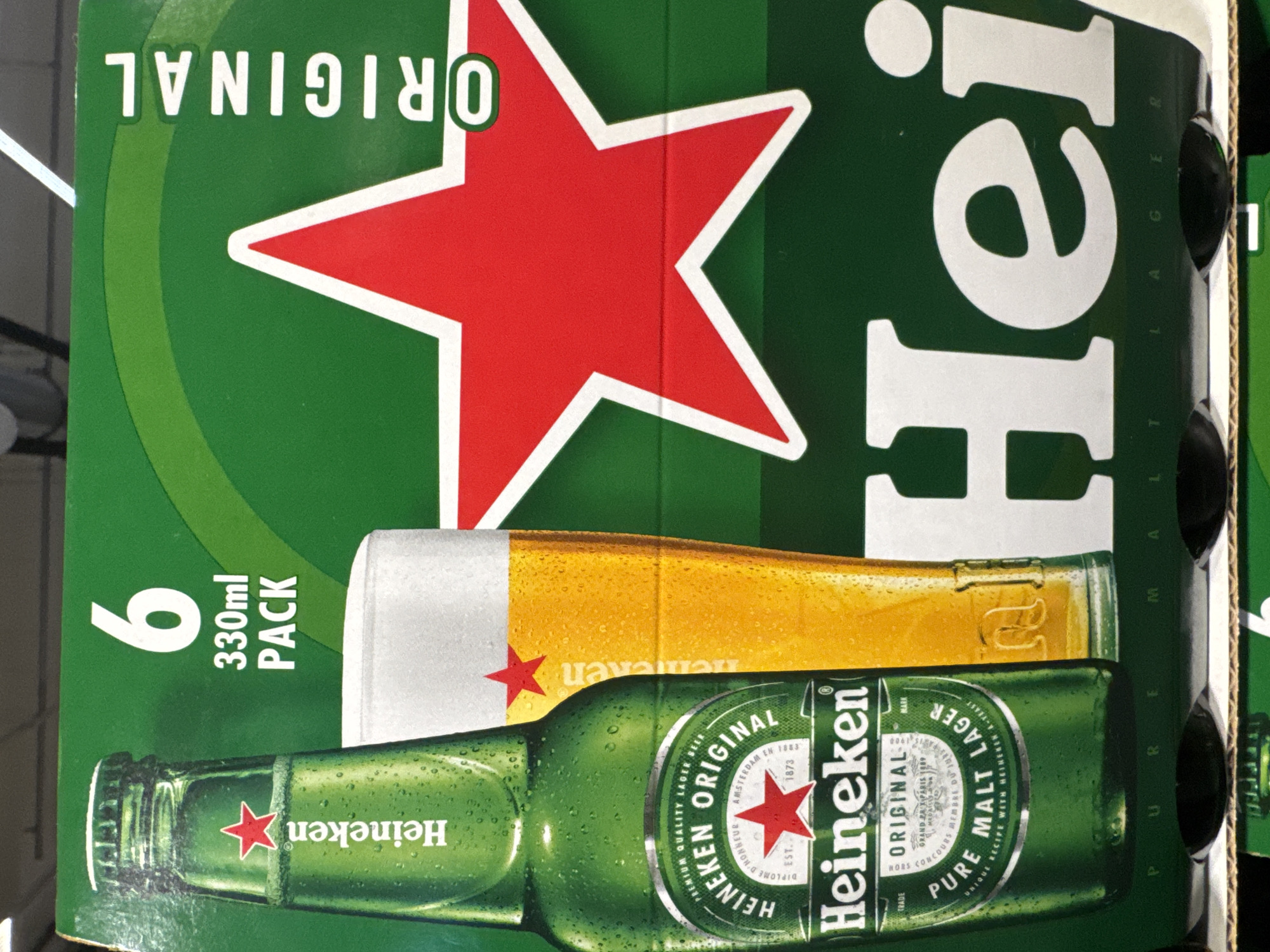 A six-pack of Heineken Original beer is displayed against a green background. The packaging features a large red star with a white outline, characteristic of the Heineken brand. One bottle of Heineken and a glass of beer are visible on the left side of the packaging, which also includes text such as "6 330ml PACK" and "ORIGINAL". The bottom of the packaging shows the text "HEI" in large white letters, partially obscured. The scene appears to be a retail environment, possibly a supermarket or liquor store.