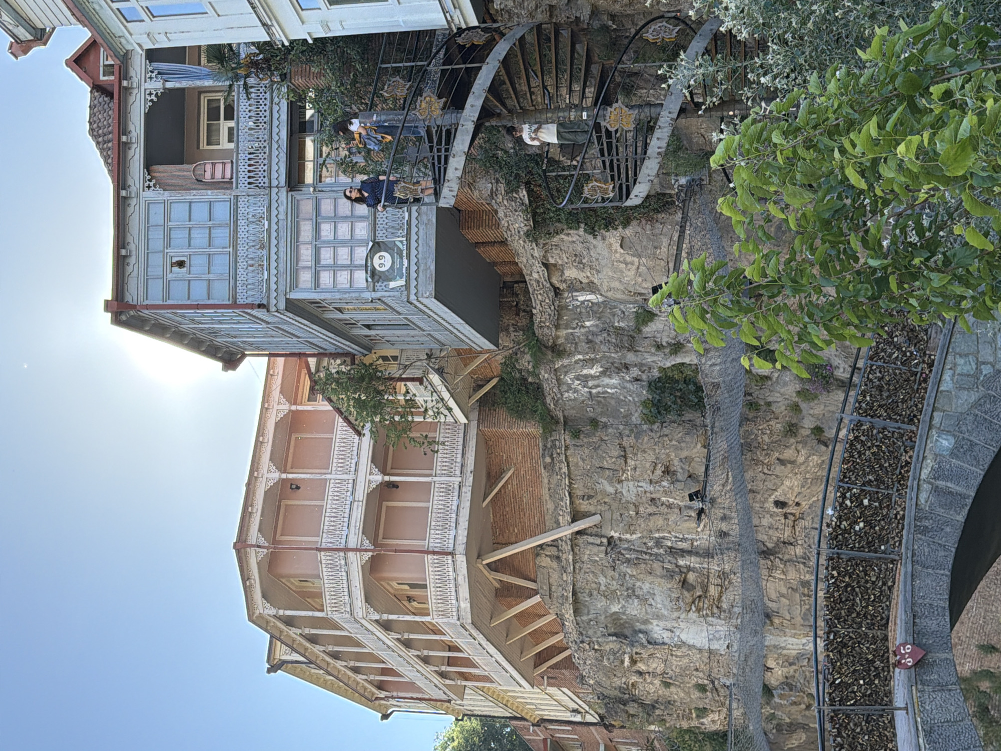 The image depicts an unusual architectural composition in Tbilisi, Georgia. Several multi-story buildings with ornate, wooden balconies are precariously attached to a steep cliff face. The buildings are painted in light hues - pink, light blue. A winding metal spiral staircase is built against the cliff, connecting the buildings. Two people are standing on the staircase. One person is wearing a white shirt and light-colored pants and another is wearing a blue dress. A third person, wearing sunglasses, is seen on a balcony. The cliff is covered with some vegetation. At the bottom right, a bridge is visible, its sides covered with numerous love locks. A safety net is installed on the cliff face, to prevent rocks from falling. The sky is clear. The sun is shining brightly, causing glare in the upper left corner.