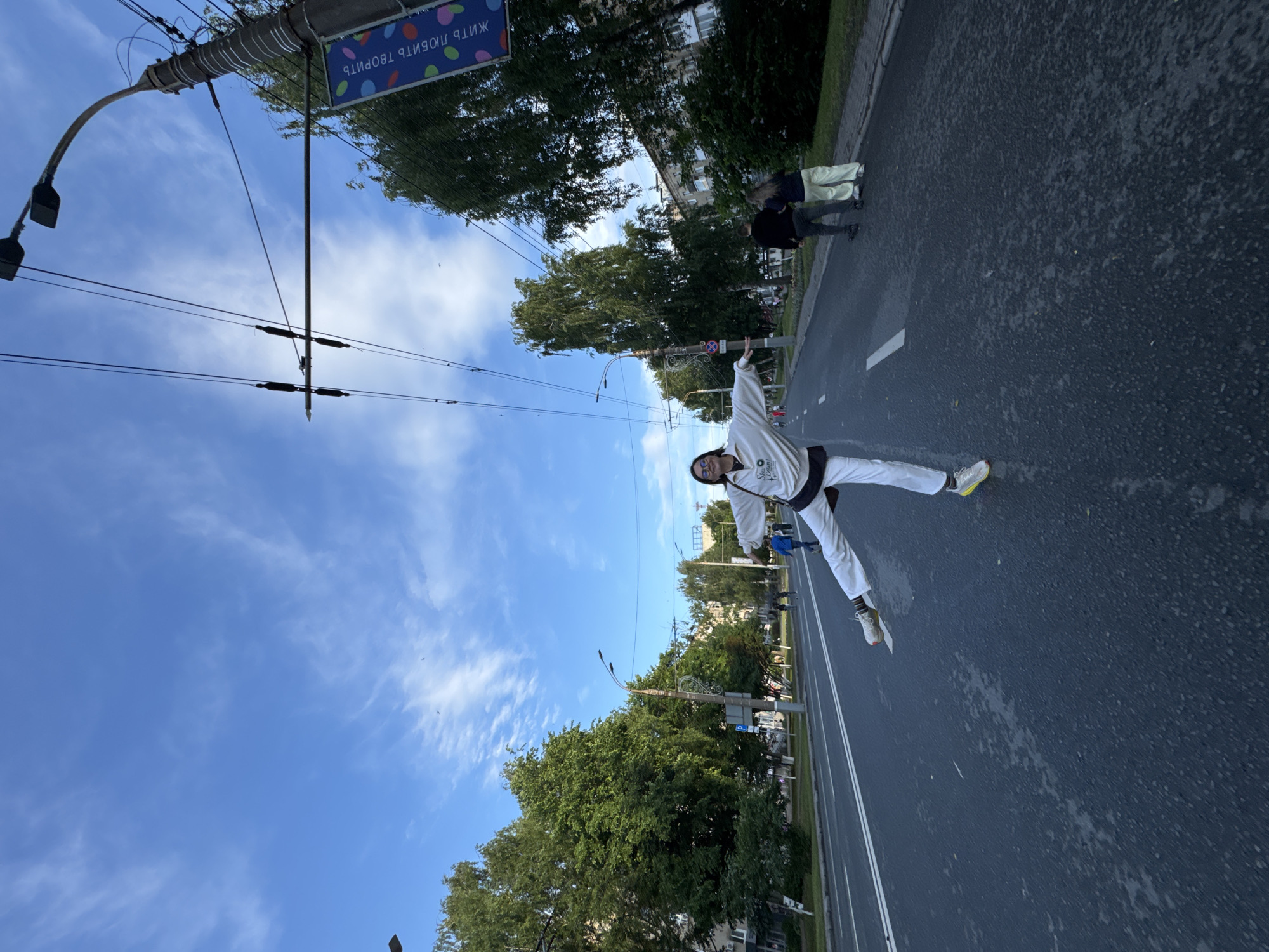 In Izhevsk, Russia, a young woman with glasses and dark hair, wearing a white hoodie and white pants, is captured mid-stride on a wide, asphalt street, her left leg extended horizontally and her right leg bent. The street is lined with trees and buildings, suggesting an urban environment. Overhead, a network of trolleybus wires and a lamppost are visible against a blue sky with scattered white clouds. A banner displaying colorful circles and Russian text, "ЖИТЬ ЛЮБИТЬ ТВОРИТЬ" (To Live, To Love, To Create), hangs from the lamppost. Two individuals, a man and a woman, walk away from the camera on the right side of the street, while other pedestrians are seen in the background on the sidewalk. The scene appears to be a public space in Izhevsk, possibly during daylight hours.
