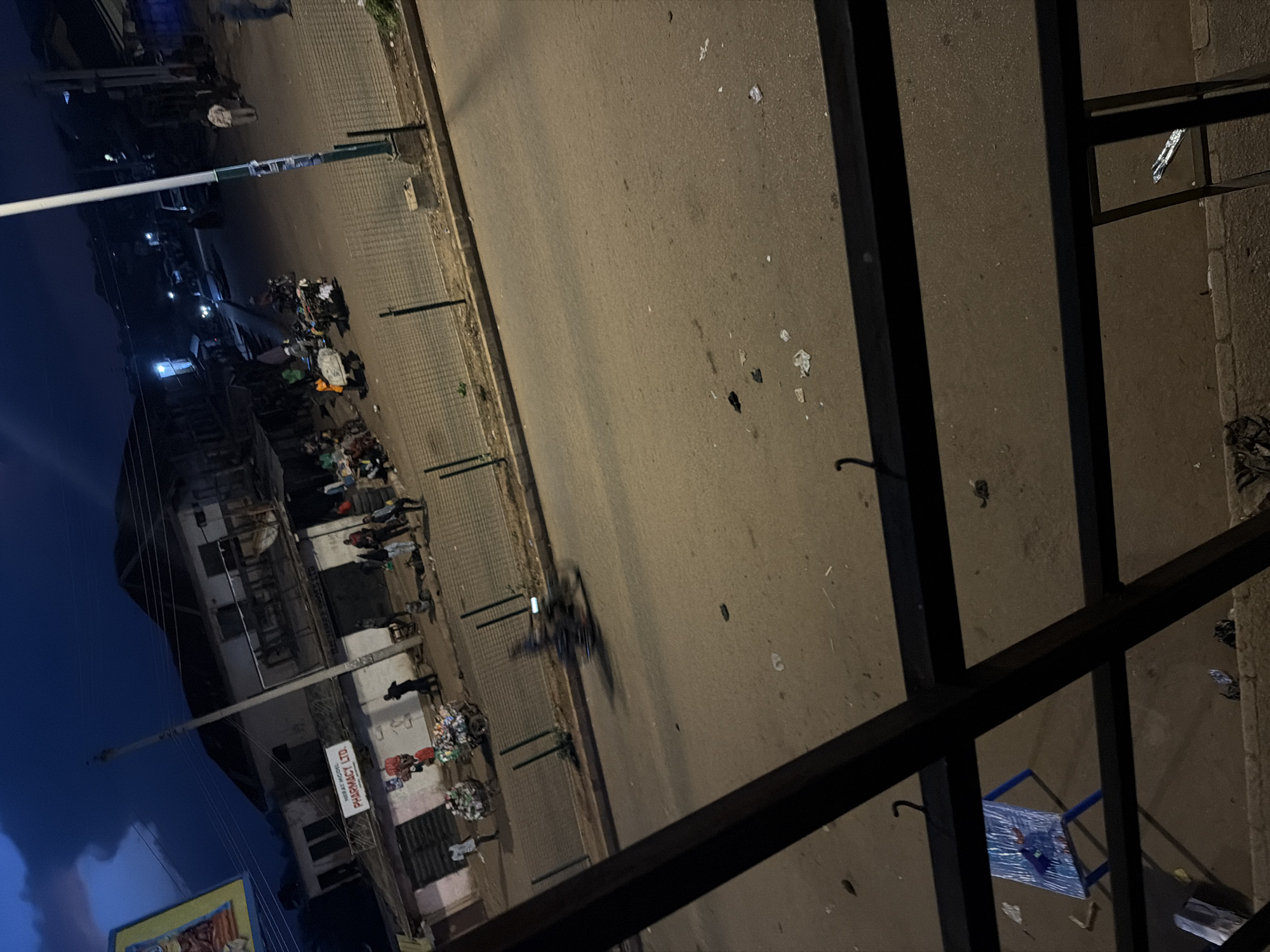 The image captures a street scene at dusk or night in Bukuru, Nigeria. A paved road occupies the foreground, littered with scattered debris including plastic bags and other refuse. A black metal railing runs across the lower portion of the frame, partially obscuring a small blue table with a colorful cartoon design. In the mid-ground, several individuals are visible, some standing near storefronts and others appearing to push carts laden with goods. A motorcycle, blurred by motion, is captured in the process of crossing the street. Across the road, a row of buildings lines the street, with a sign for "MERAT MODEL PHARMACY LTD." prominently displayed on one of them. The sky above is dark blue with hints of clouds, and streetlights illuminate parts of the scene, casting long shadows. The overall atmosphere suggests a bustling marketplace or commercial area during its closing hours.