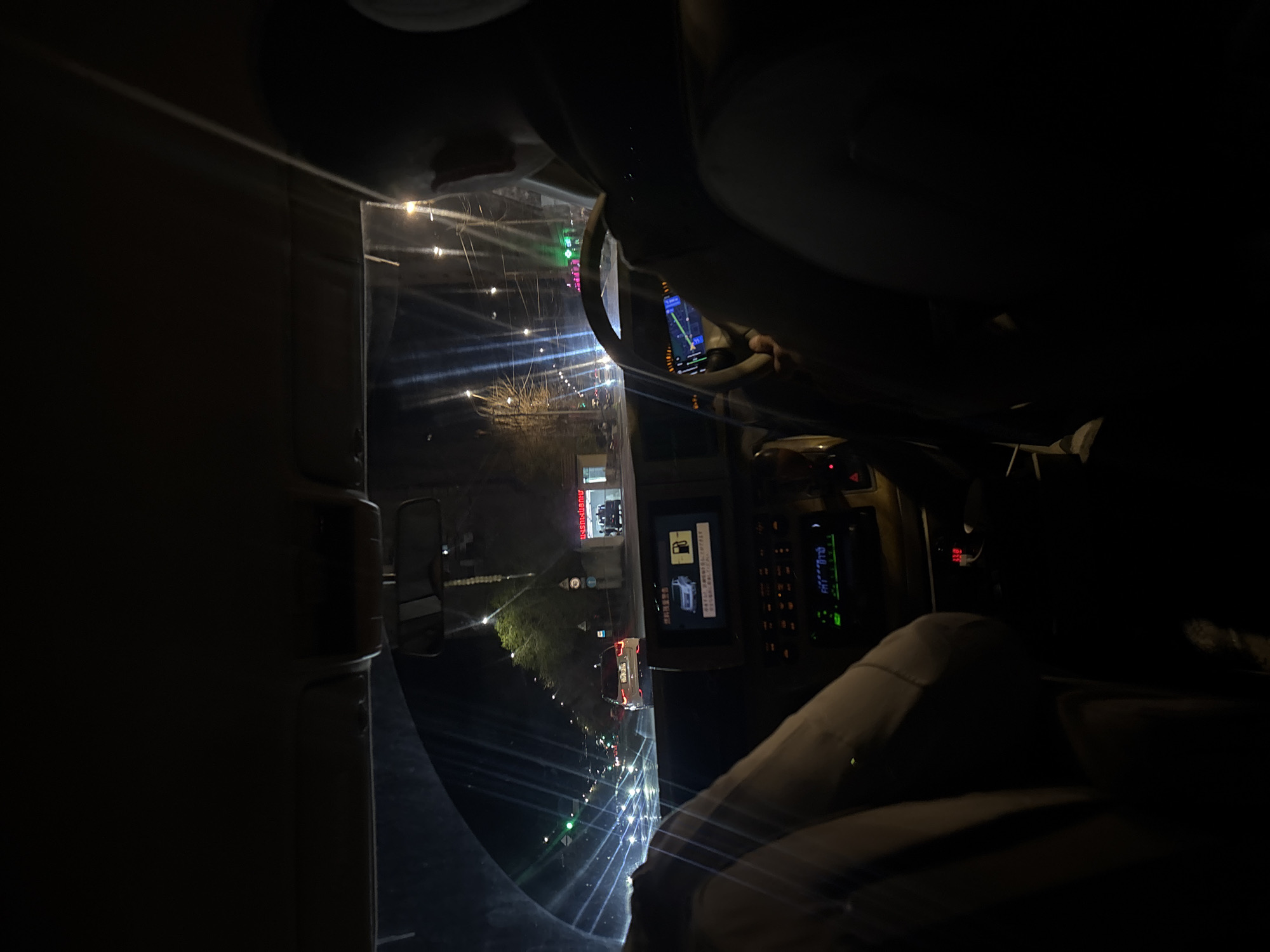 The image depicts a view from inside a vehicle, likely a truck, at night. Through the windshield, a street scene unfolds, illuminated by streetlights and the headlights of oncoming traffic. Buildings are visible along the roadside, including one with illuminated signage in Cyrillic script, suggesting a location in Yerevan, Armenia. Inside the vehicle, the dashboard is visible, with various controls and a digital display showing a diagram of a vehicle refueling. The steering wheel is partially visible. The scene is dark, with light sources concentrated on the street and dashboard.