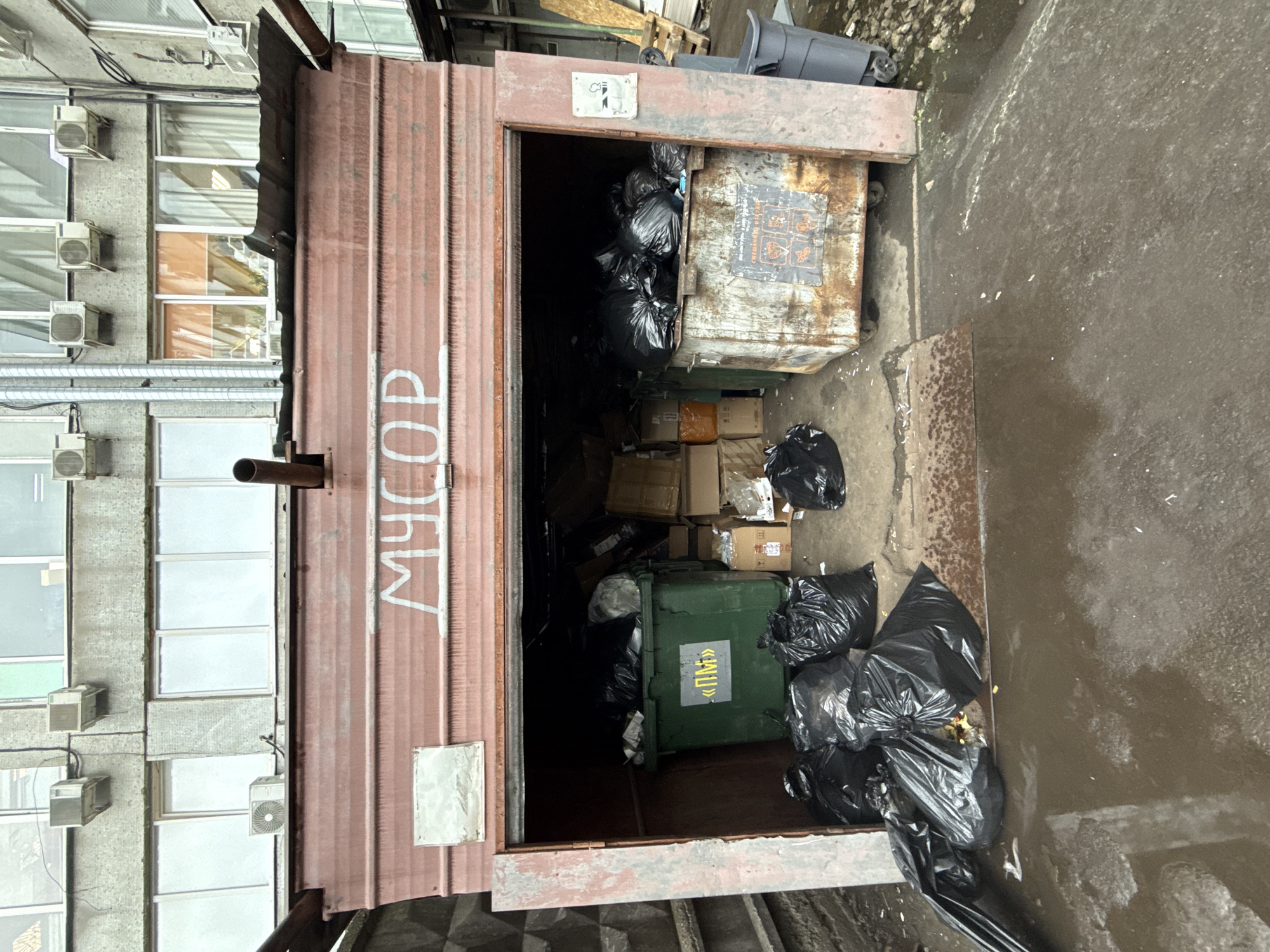 The image shows a trash collection area in Dolgoprudny, Russia. The trash is located in an open-fronted, red metal container with the Russian word for "garbage" ("МУСОР") written vertically on the side. The container is full of black plastic trash bags, cardboard boxes, and a green recycling bin. A large, rusted metal dumpster is positioned towards the rear of the enclosure, also filled with trash bags. Outside the container, more black plastic bags are piled up on the ground. The area is paved with dark concrete. A building with multiple windows and air conditioning units is to the left of the trash container.