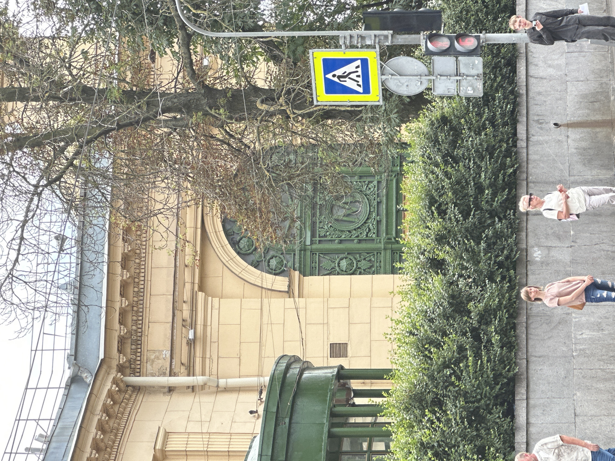 This image depicts a street scene in Saint Petersburg, Russia, likely near a public building. In the foreground, a dense green hedge forms a barrier along a grey stone wall. Several people are visible standing along this wall. To the right, a man in a dark shirt and trousers holds a phone. Next to him, a woman in light-colored clothing with a pink backpack stands and looks away. Further left, a woman in a pink t-shirt and ripped jeans stands with her arms crossed, carrying a brown bag. Another person is partially visible to the far left. Behind the hedge and wall, a tall, bare-branched tree stands in front of a building with light yellow facade. The building features decorative architectural elements, including an arched entrance with an ornate green metal gate. A green, rounded structure, possibly an entrance canopy or kiosk, is visible to the left of the building. A pedestrian crossing sign and a traffic light, showing red for vehicles, are positioned near the tree and the building. The sky is overcast.