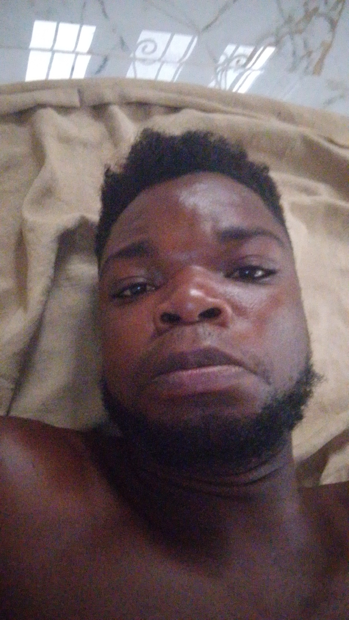 A close-up, upward-angled photo shows a dark-skinned man with a dark beard and hair against a light tan fabric background. His facial expression is neutral. Above him, a ceiling tile pattern is partially visible, suggesting an indoor location. The specific location in Enugwu-Ukwu, Nigeria, cannot be determined from this image.