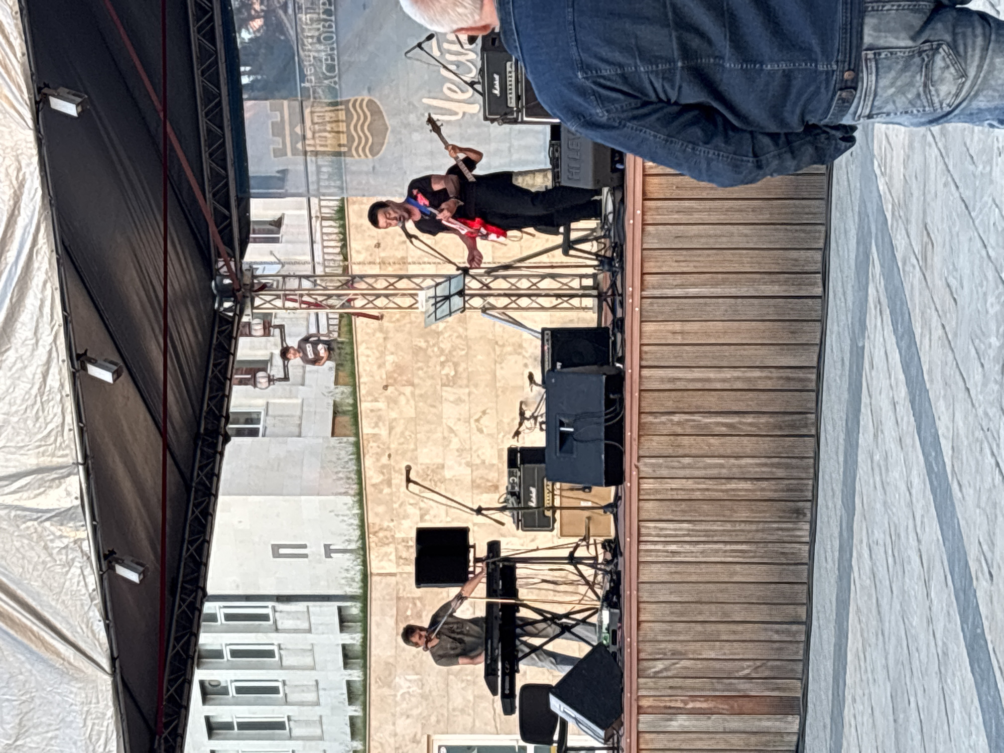 A band is performing outdoors in Asenovgrad, Bulgaria. A guitarist in black clothes with a red guitar plays on a wooden stage, and a keyboardist sits at a keyboard lower on the stage. Stage lighting and sound equipment are visible. An audience member in a blue denim jacket stands in the foreground, partially obscuring the view. The backdrop behind the stage features an image resembling a city scene and text, possibly related to Asenovgrad's identity. Buildings surround the location of the performance.
