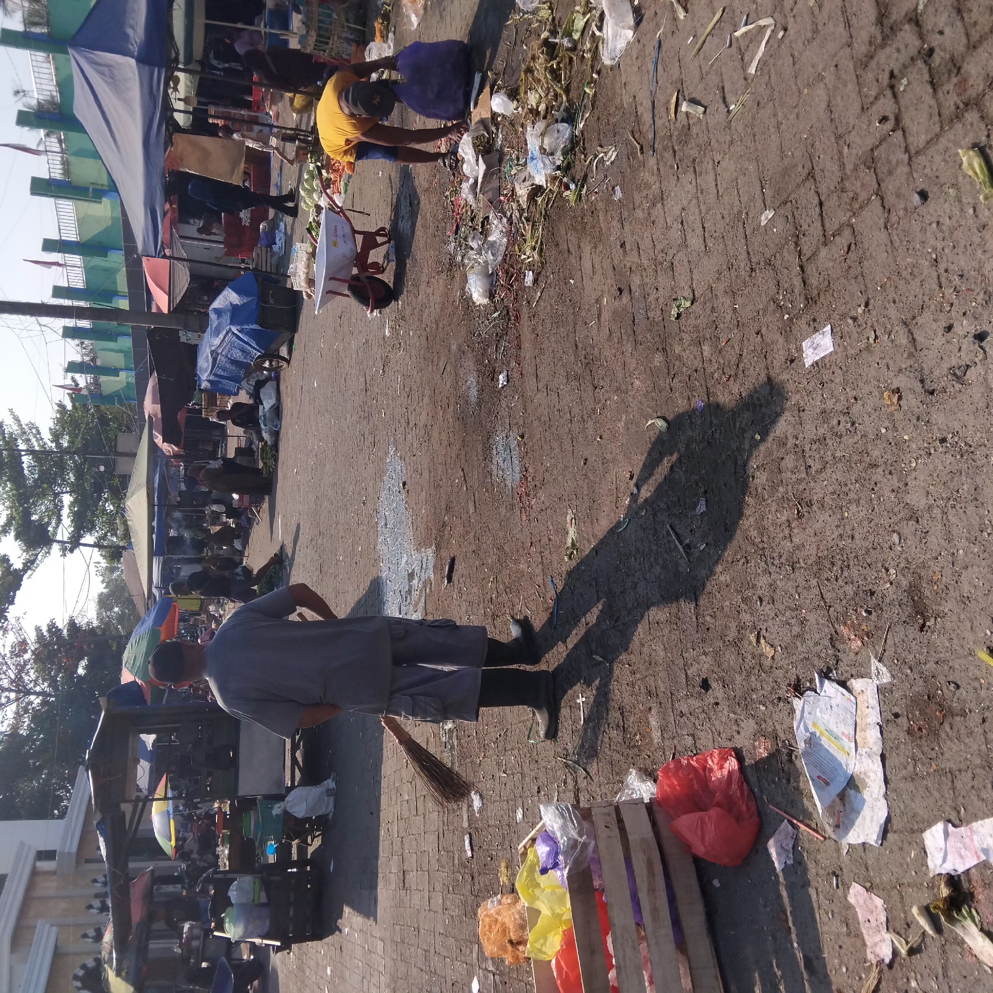 A street market scene in Banjarbaru, Indonesia, shows a person in grey shorts and dark boots sweeping the ground with a broom. Scattered trash, including plastic bags and food debris, litters the brick pavement. Several market stalls with colorful umbrellas are visible in the background, along with other individuals. A person in a yellow shirt is seen collecting trash in a purple bag near a wheelbarrow. The ground is uneven and dusty, suggesting a busy commercial area.