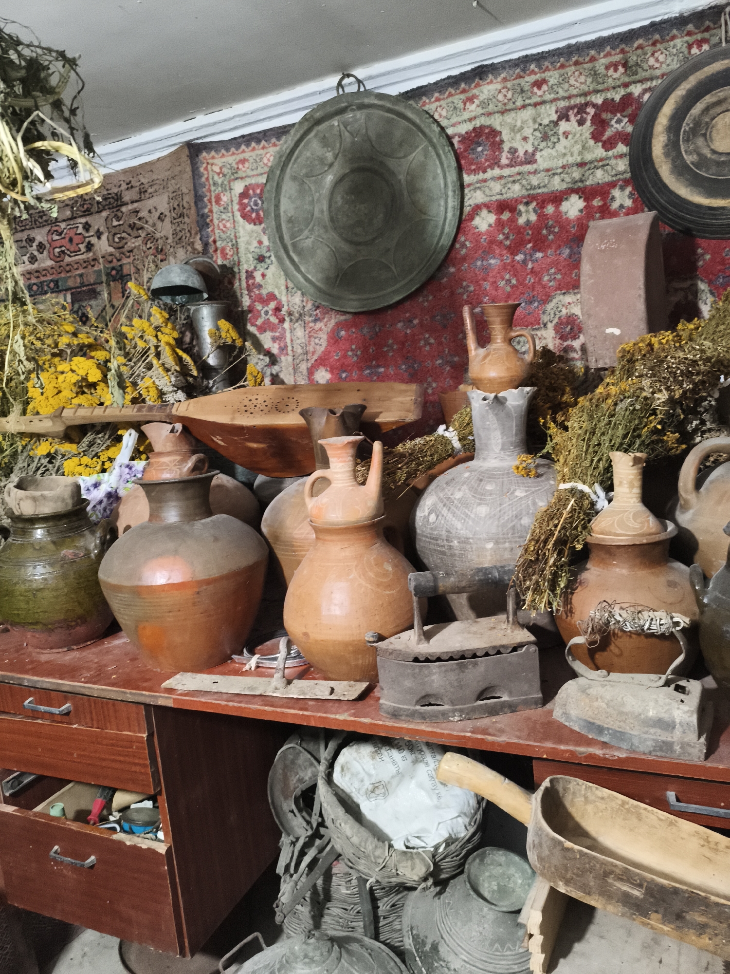 The image depicts a cluttered room that appears to be a storage space or a workshop. The scene is filled with numerous antique items, primarily clay pots and jugs of various sizes and designs, some with intricate patterns. There are also several old-fashioned flat irons, a wooden instrument that resembles a lute or a dulcimer, and bundles of dried herbs or flowers. Two large, decorative rugs with intricate patterns hang on the wall behind the displayed items. The items are arranged on a wooden shelf and on the floor. The lighting is dim, casting shadows and highlighting the aged textures of the objects. The overall impression is one of a collection of historical artifacts, possibly in a rural or historical setting. The mention of Irgali, Russia, suggests this location is the specific setting for this collection of items.