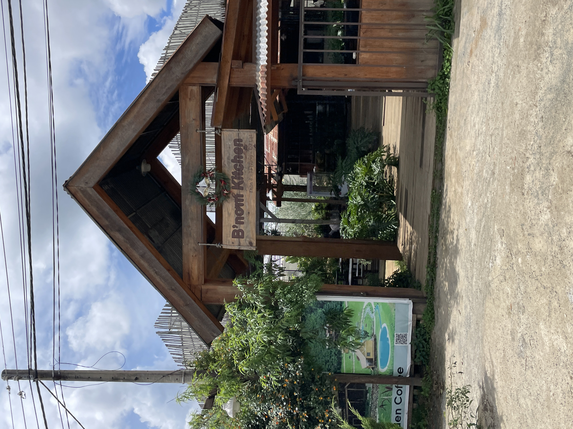 A wooden building with a pitched roof, decorated with a Christmas wreath, is pictured in Thôn 4, Vietnam. The sign above the entrance reads "B'nom Kitchen". A wooden walkway leads into the building, which appears to be a restaurant or cafe. Lush greenery surrounds the structure, with trees and plants visible in the foreground and background. A cloudy sky with patches of blue can be seen above. To the left of the building, a poster displays an image of a house by a pond. Electrical wires and a utility pole are also present in the scene. The ground in front of the building is a mixture of dirt and gravel.