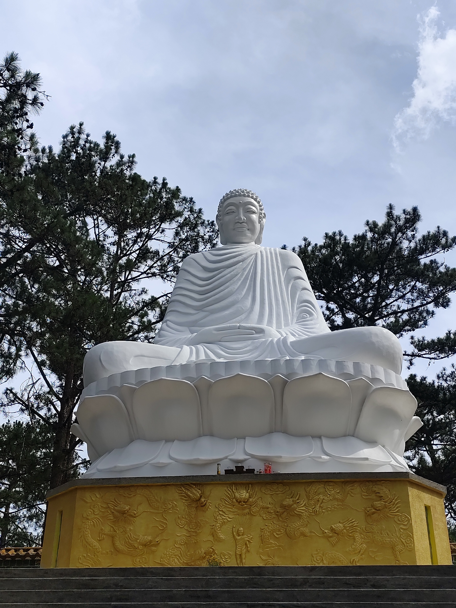 A white Buddha statue sits atop a yellow, ornate pedestal in Da Lat, Vietnam. The statue is positioned in front of dark green pine trees against a cloudy sky. The pedestal features intricate dragon carvings. A few offerings are placed at the base of the statue. Stone steps lead up to the statue.