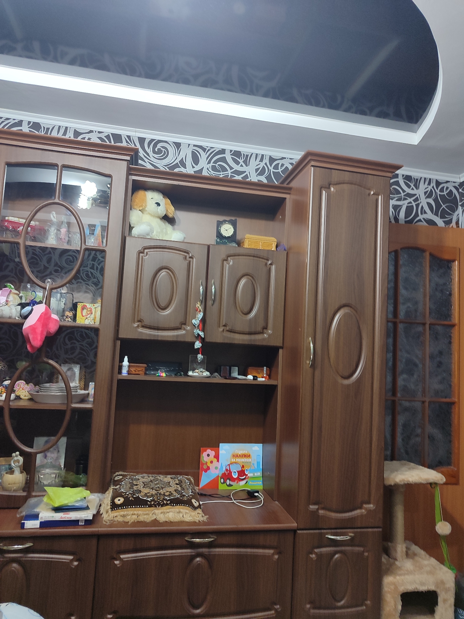 The image shows a living room with a large, wooden wall unit. The wall unit has several shelves and cabinets, filled with various items. On the top shelf of the right side of the unit, a stuffed dog is placed. Below it, there are two cabinet doors. The left side of the unit has glass-fronted shelves, displaying decorative items and some books. A plush toy in pink and red is hanging from a shelf on the left side. In front of the wall unit, a dark brown cabinet with drawers is visible. On top of this cabinet, there are several items including books, a patterned pillow, and two greeting cards. The room has patterned wallpaper and a dark, curved ceiling. A cat tree with a scratching post and platforms is located to the right of the wall unit, partially visible. The overall impression is of a well-decorated but lived-in space. The scene is likely located in an apartment or house in Mykolaiv, Ukraine, given the presence of Cyrillic text on the books and the general style of the furniture.