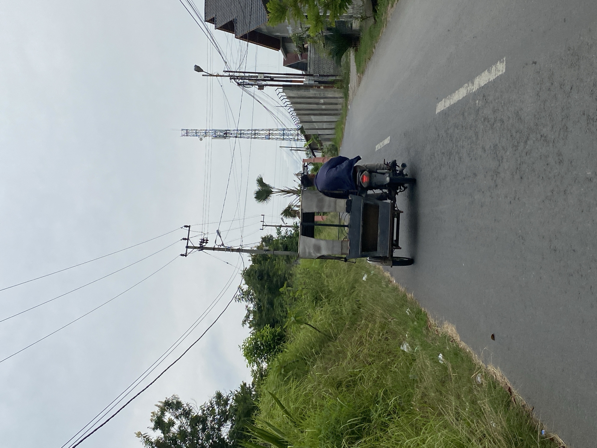 An overcast day in Lhokseumawe, Indonesia, shows a person riding a modified motorcycle, possibly a becak or a tricycle taxi, on a paved road. The rider, wearing a dark jacket and light-colored pants, is in the center of the frame, facing away from the camera. The motorcycle is pulling a passenger carriage, which is empty. To the left of the road is a steep embankment covered in tall, green grass and some scattered debris. On the right side of the road, there is a concrete wall, a house with a dark roof, and a tall communications tower in the distance. Power lines crisscross the sky, supported by utility poles. The road has a broken white line marking its center.