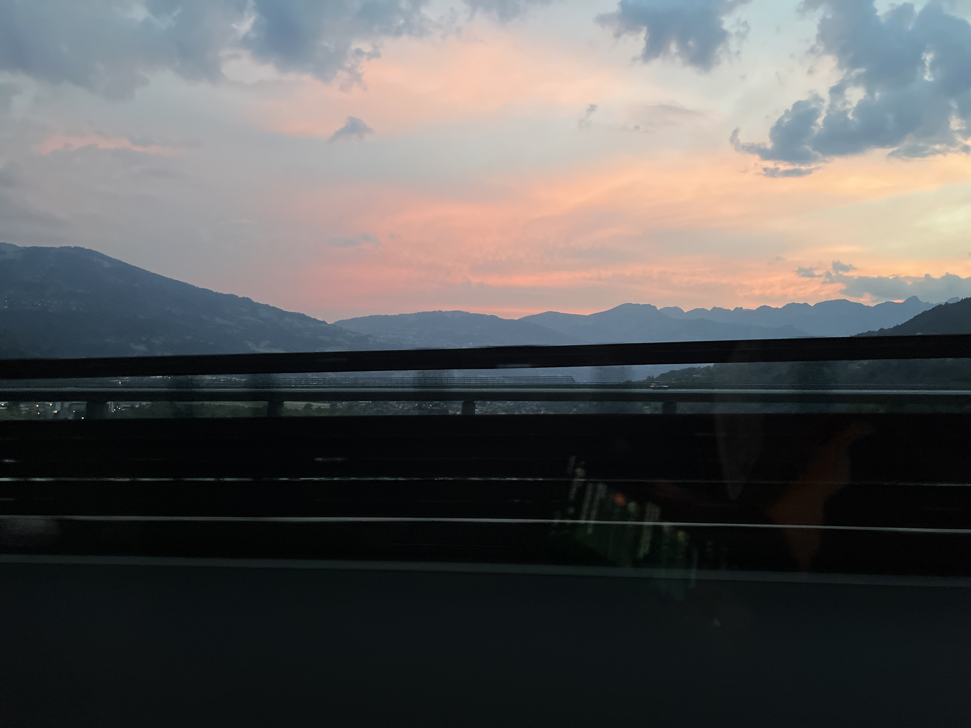 This image depicts a view from a vehicle traveling in the city of Passy, France. The scene is dominated by a twilight sky filled with a gradient of colors ranging from soft pinks and oranges near the horizon to cooler blues and grays higher up. Dark, silhouetted mountain ranges form a backdrop against the colorful sky, their contours softened by the fading light. In the midground, a series of horizontal dark structures, likely part of a highway or bridge, span across the frame. Beyond these structures, a dimly lit landscape is visible, suggesting a town or city with scattered lights. The foreground of the image is obscured by dark, reflective surfaces, possibly the windows of the vehicle, which show faint reflections of the interior and the passing scenery. The overall impression is one of a scenic drive through a mountainous region during sunset.