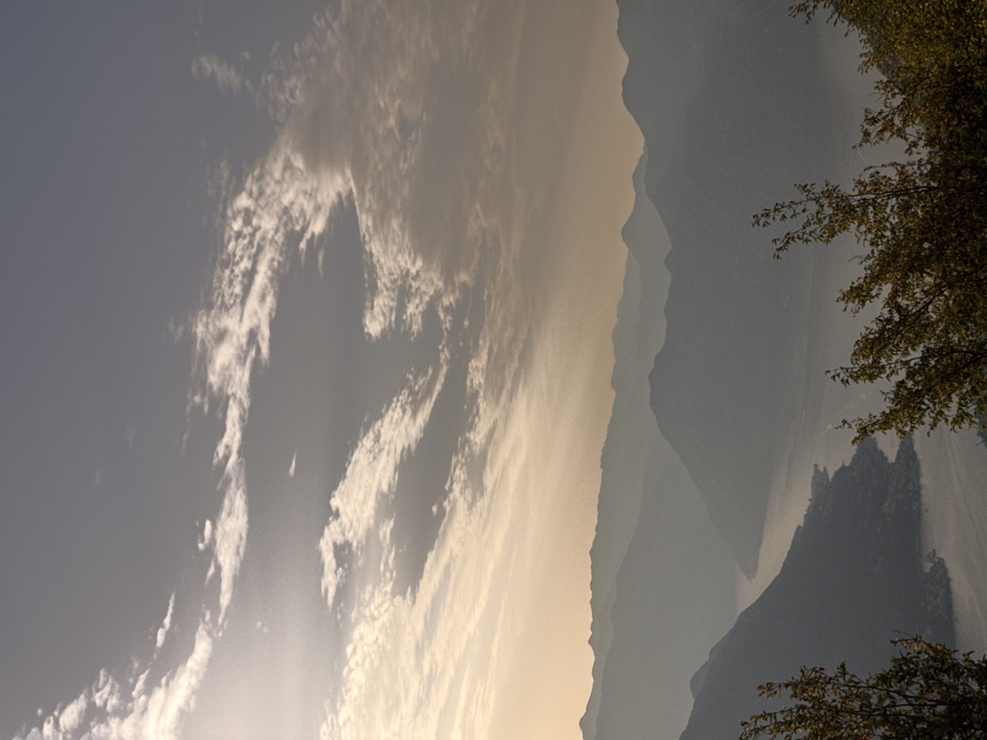 The image depicts a serene landscape of mountains and a body of water, likely a fjord, viewed from a high vantage point. The scene is bathed in the soft, diffused light of either dawn or dusk, casting a hazy glow over the layered mountain ranges that recede into the distance. Wispy clouds are scattered across the sky, adding texture and depth to the scene. In the foreground, the dark silhouettes of trees frame the lower portion of the image, their branches reaching into the view. A settlement is visible along the shoreline of the water, its lights appearing as small, indistinct specks in the gathering twilight. The overall impression is one of tranquility and natural beauty. The location is identified as Žanjev Do, Montenegro.
