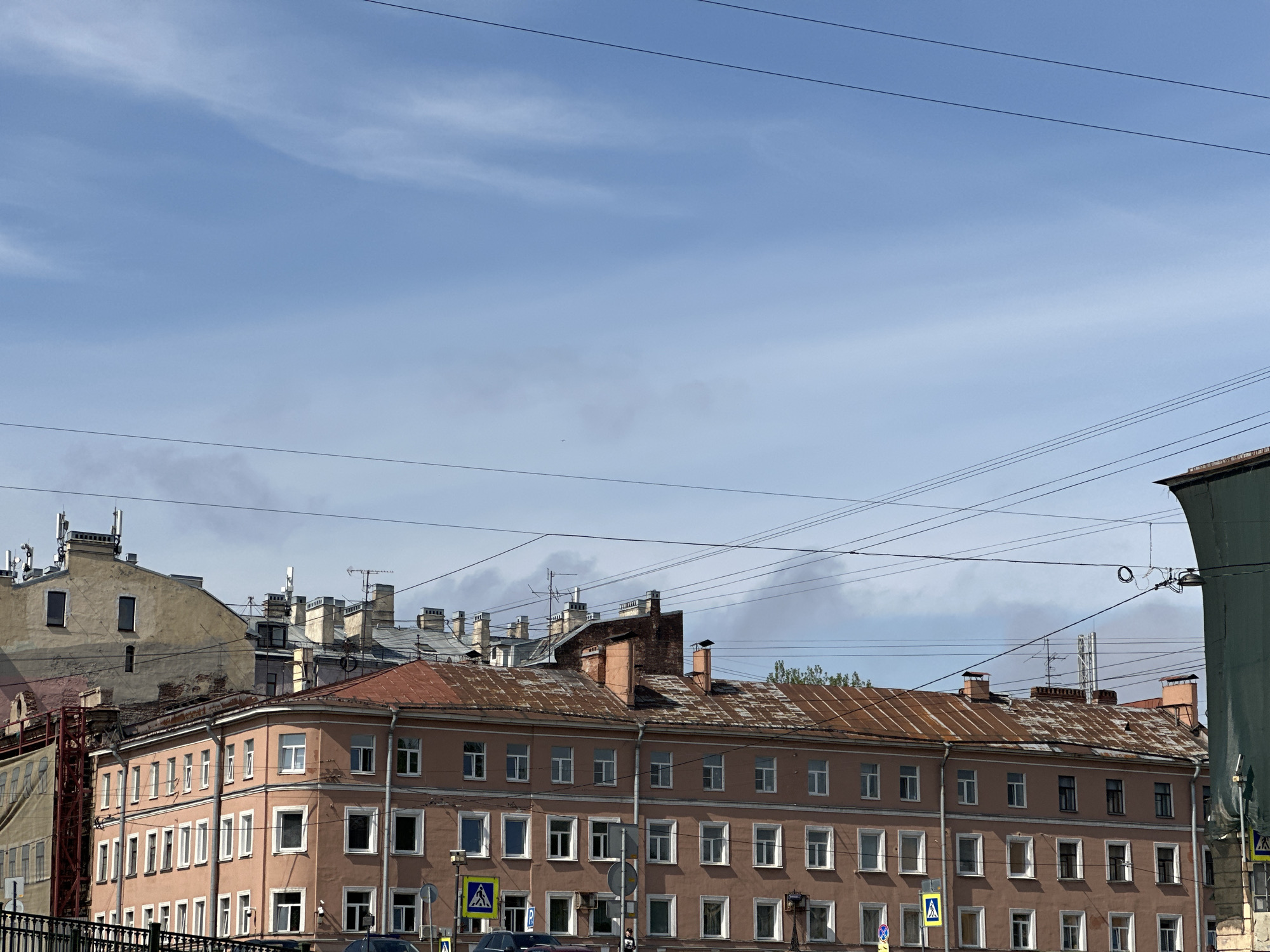 The scene depicts an urban environment in Saint Petersburg, Russia, presenting a view of buildings against a blue sky with scattered cirrus clouds. Numerous overhead electrical lines crisscross the sky. The architecture of the buildings suggests a historical urban setting. A four-story building with a reddish-brown facade occupies a significant portion of the frame. The building exhibits a repeating pattern of windows. Other structures of varying heights, materials, and conditions are visible in the background, indicating a densely populated area. Signage, including pedestrian crossing signs, is present near street level. Some cars can be seen in front of the four-story building. The overall impression is of a typical cityscape in Saint Petersburg.