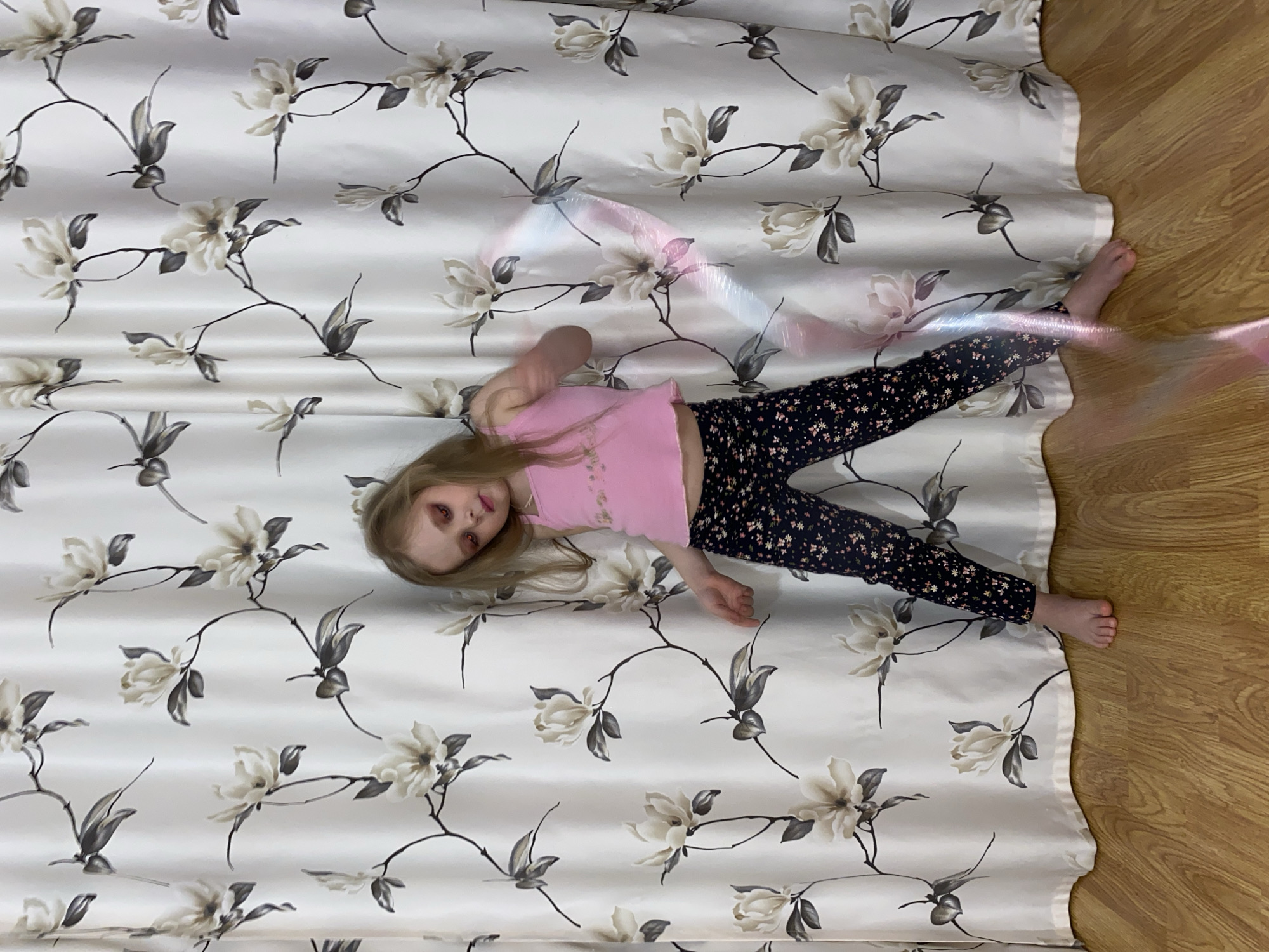 A young girl lies on her back on floral-patterned fabric that extends onto a wooden floor. She wears a pink top and dark leggings with a small floral pattern. A blurred light effect obscures parts of her body and the fabric. The scene appears to be indoors. It's unknown if the image was taken in Votkinsk, Russia.