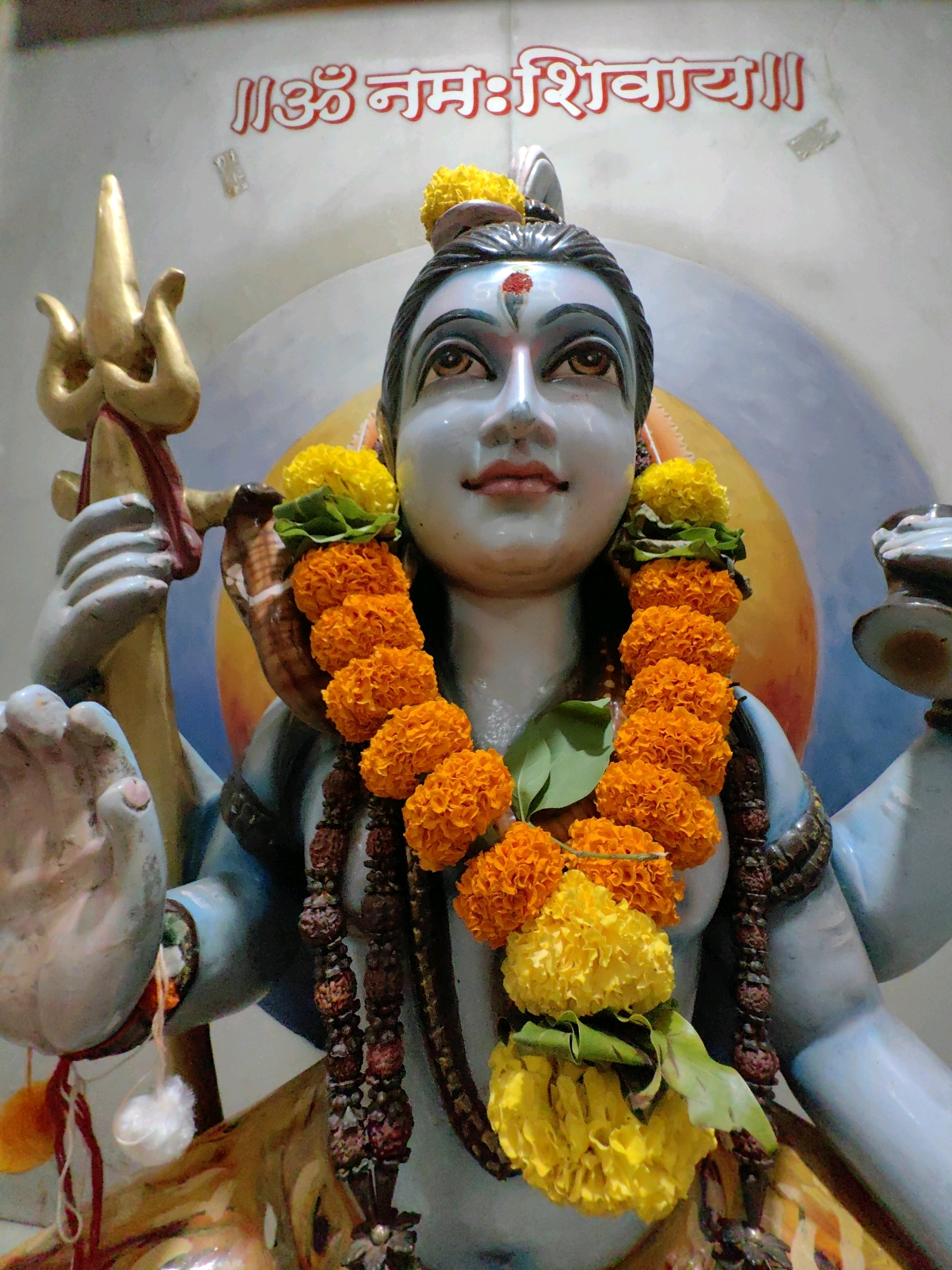 The image is a close-up, eye-level shot of a statue of the Hindu deity Shiva. The statue is blue-skinned and adorned with garlands of orange marigolds and yellow flowers. It wears a rudraksha bead necklace and a red tika on its forehead. Shiva holds a golden trident in its left hand. Behind the statue is a blurred background with a large, orange and blue circular element. Above the statue, text in red Devanagari script is visible. The statue appears to be located indoors, possibly in a temple or shrine. The text above the statue reads "ॐ नमः शिवाय" which is a Hindu mantra dedicated to Lord Shiva. The overall impression is one of religious devotion and artistic representation. The image does not contain any identifiable Mumbai landmarks or context to place it specifically within the city of Mumbai, India.