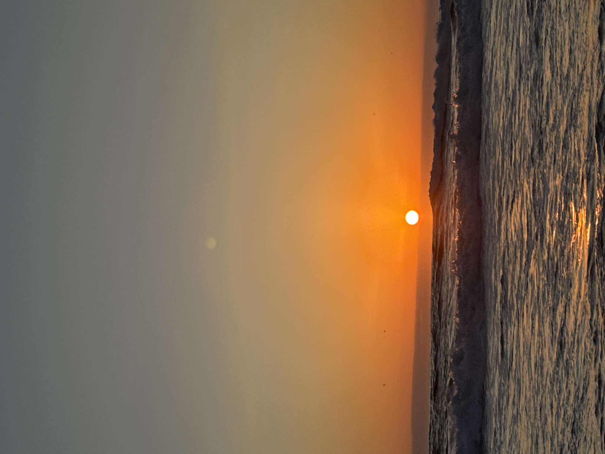 A vertical shot captures a beach scene at sunset. The sky is a gradient of grey to orange as it meets the horizon, where the sun appears as a bright, small orb. Next to the sky is the ocean, with visible waves. The light from the setting sun reflects off the waves, creating a golden hue.